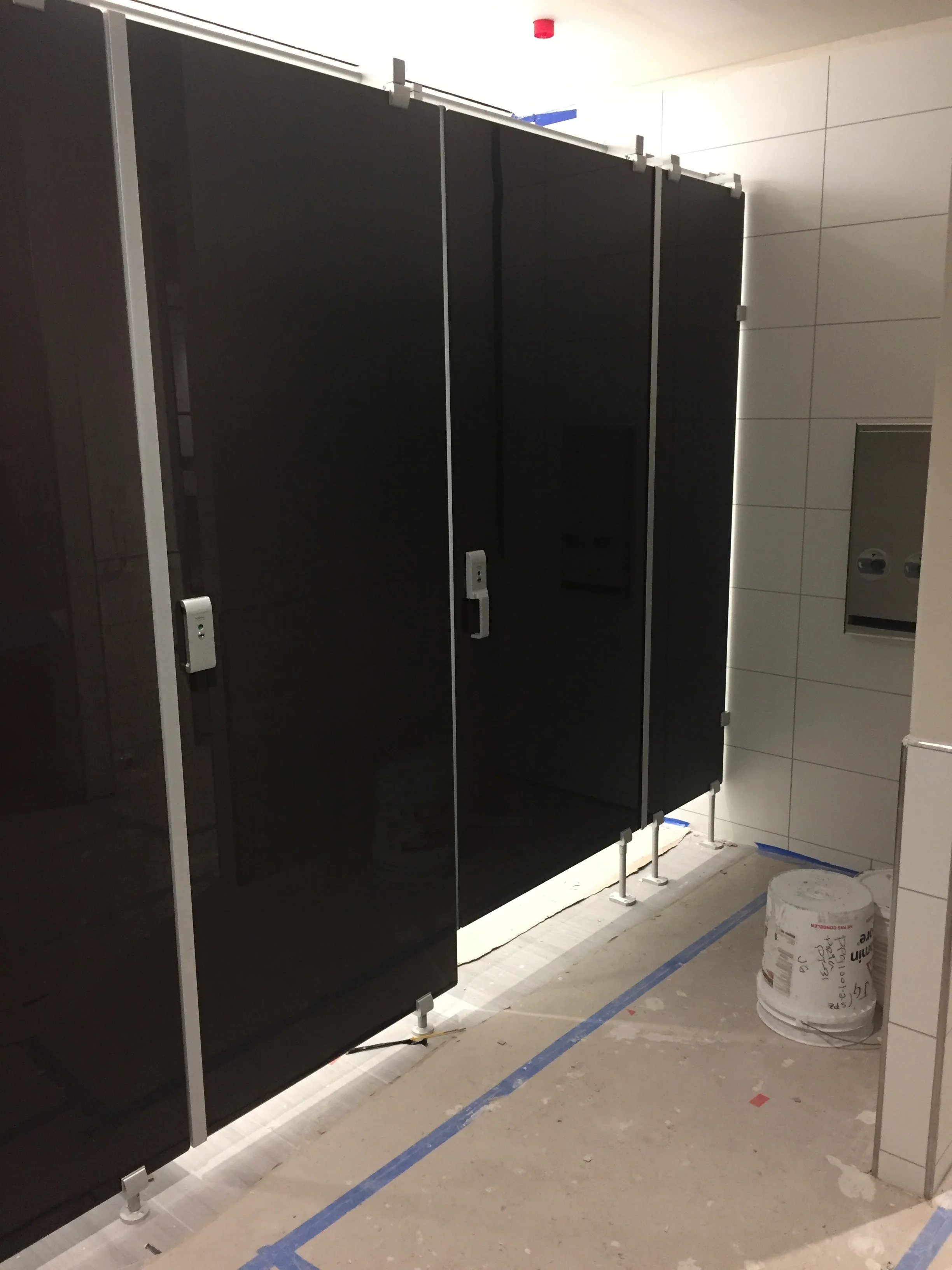 Public restroom with black partition stalls, white tiled walls, and construction materials on the floor.