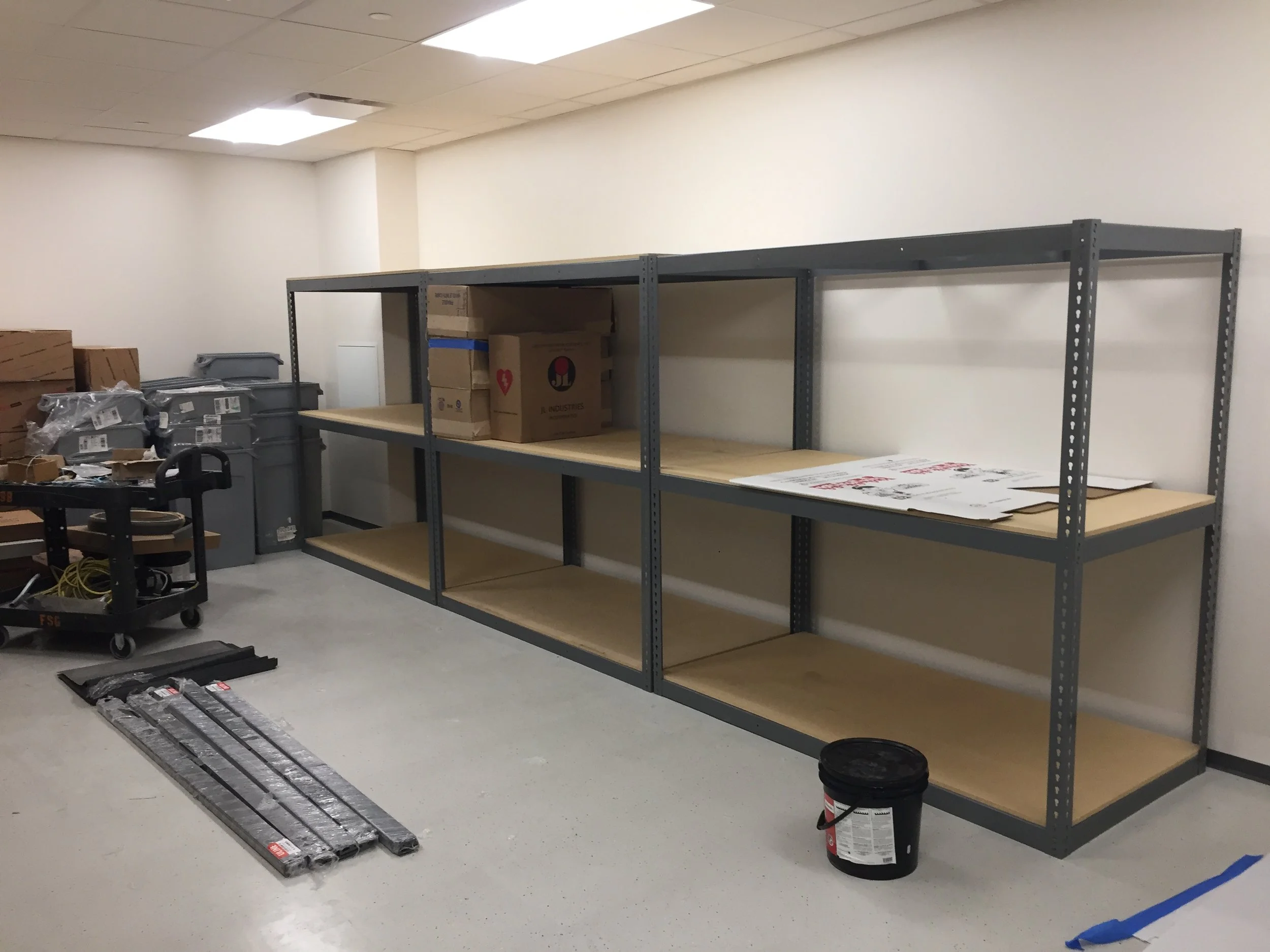 Empty storage room with metal shelves, some boxes, plastic bins, and packaging supplies on the floor.