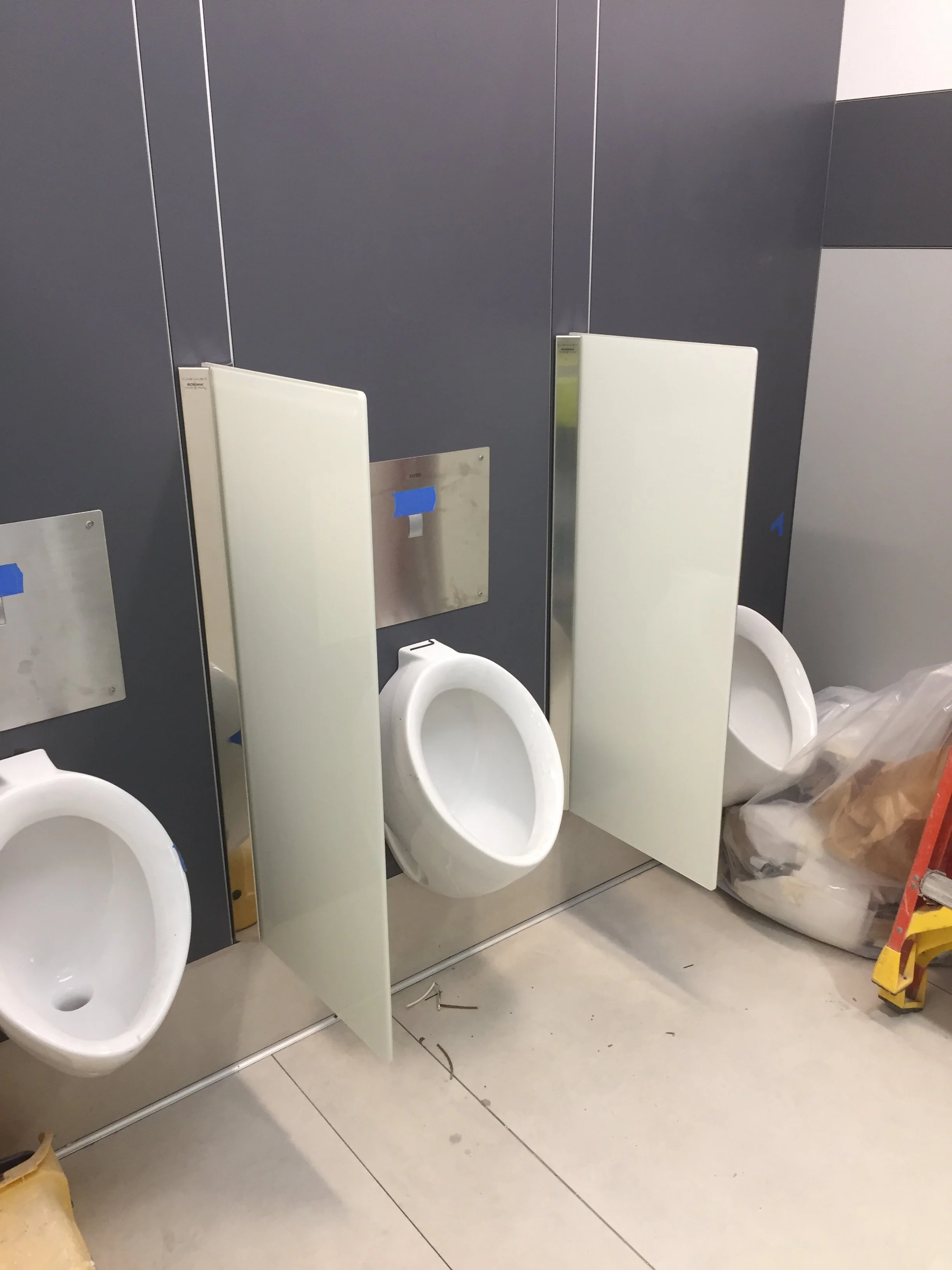 Public restroom with two urinals separated by privacy dividers, a partially visible trash bag on the right, blue painter's tape on a metal wall plate, and tiled floor with some debris.