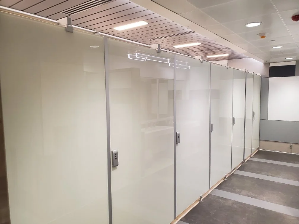 Empty glass or acrylic partitioned cubicles in a modern office or restroom setting, with metal locks on each door and a grey floor with white lines.