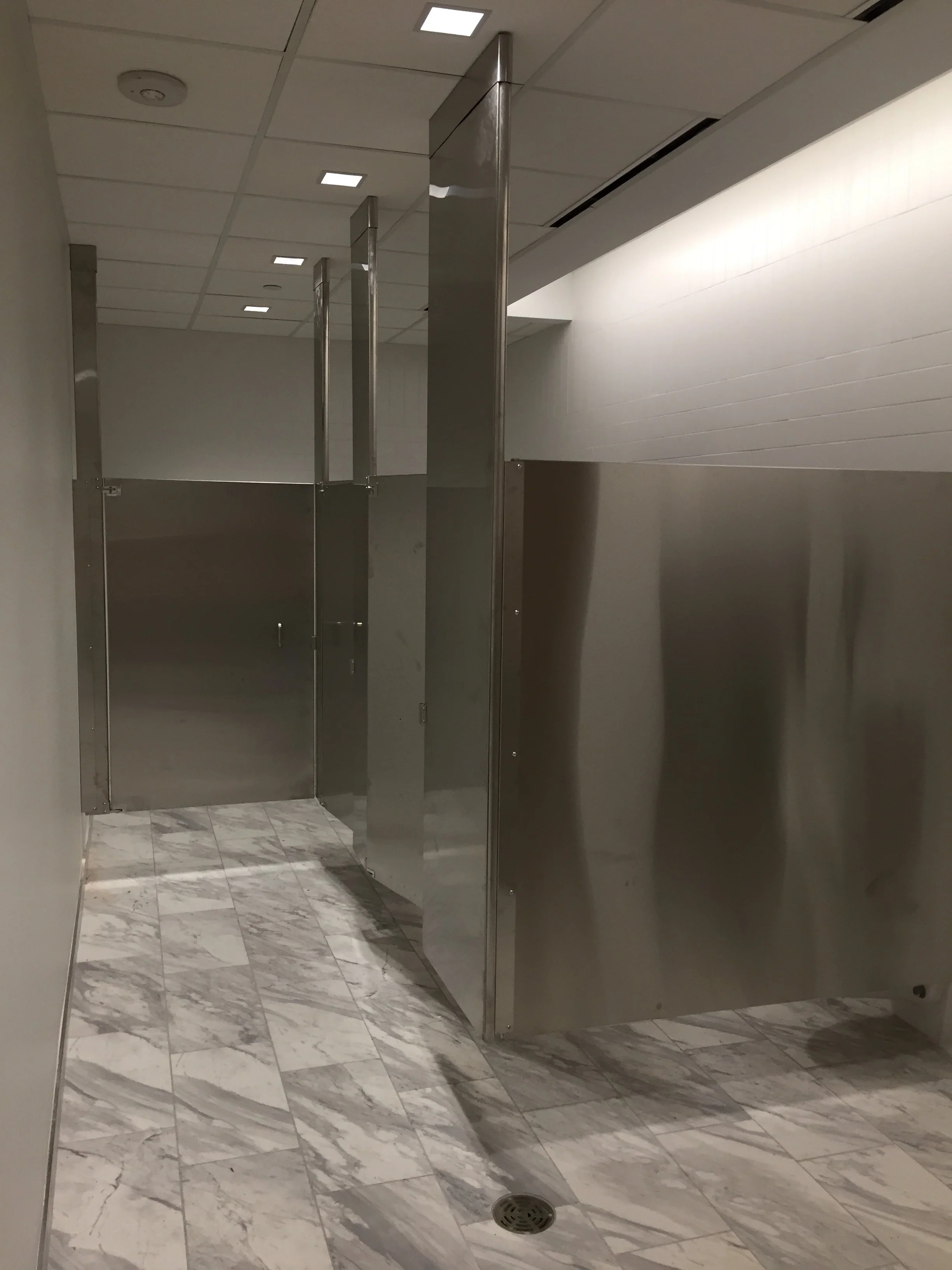 Public restroom with metal stalls, marble floor, and white tiled wall.