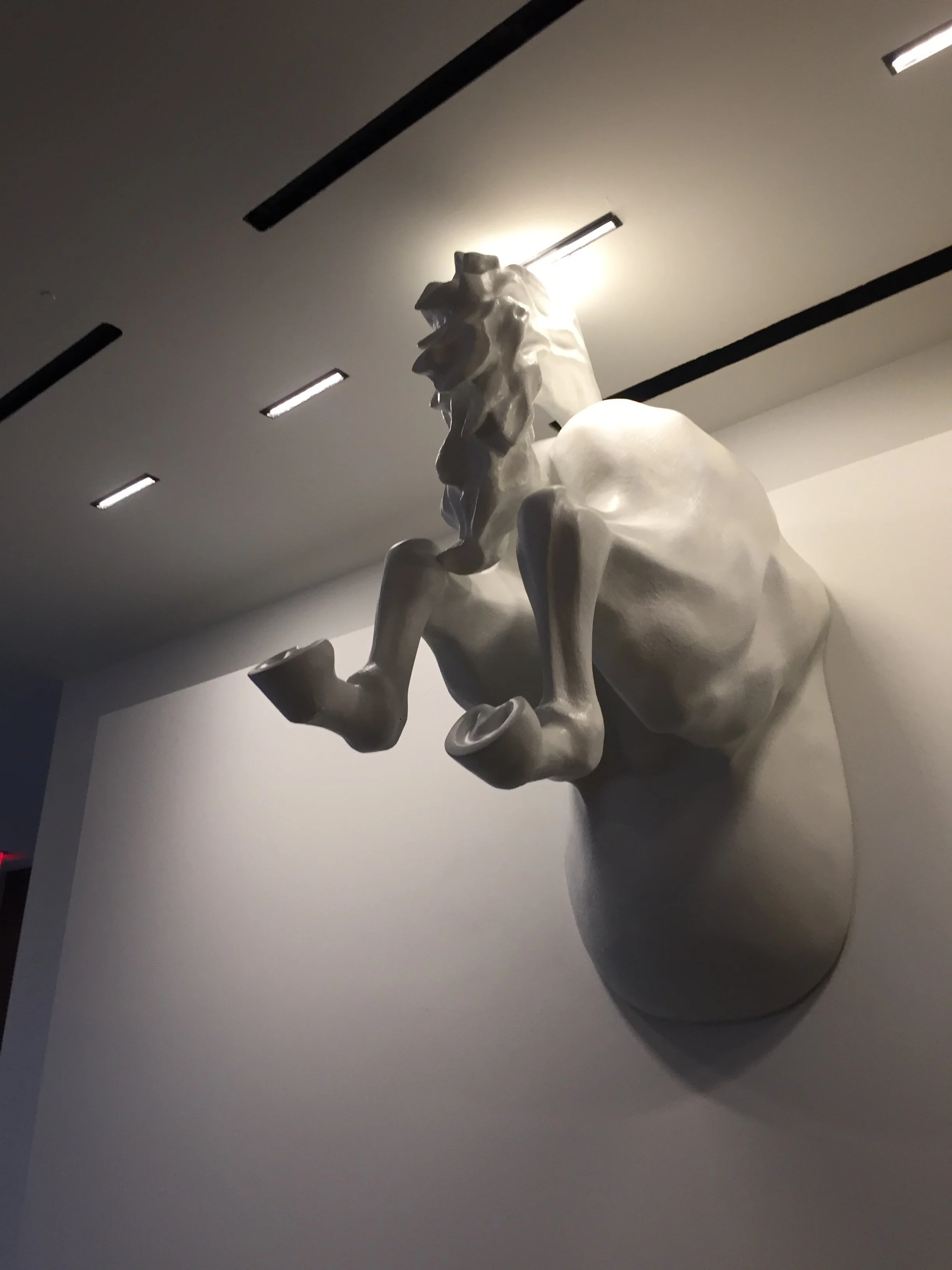 White sculpture of a lion with a raised paw, mounted on a round pedestal in an art gallery with overhead lighting.