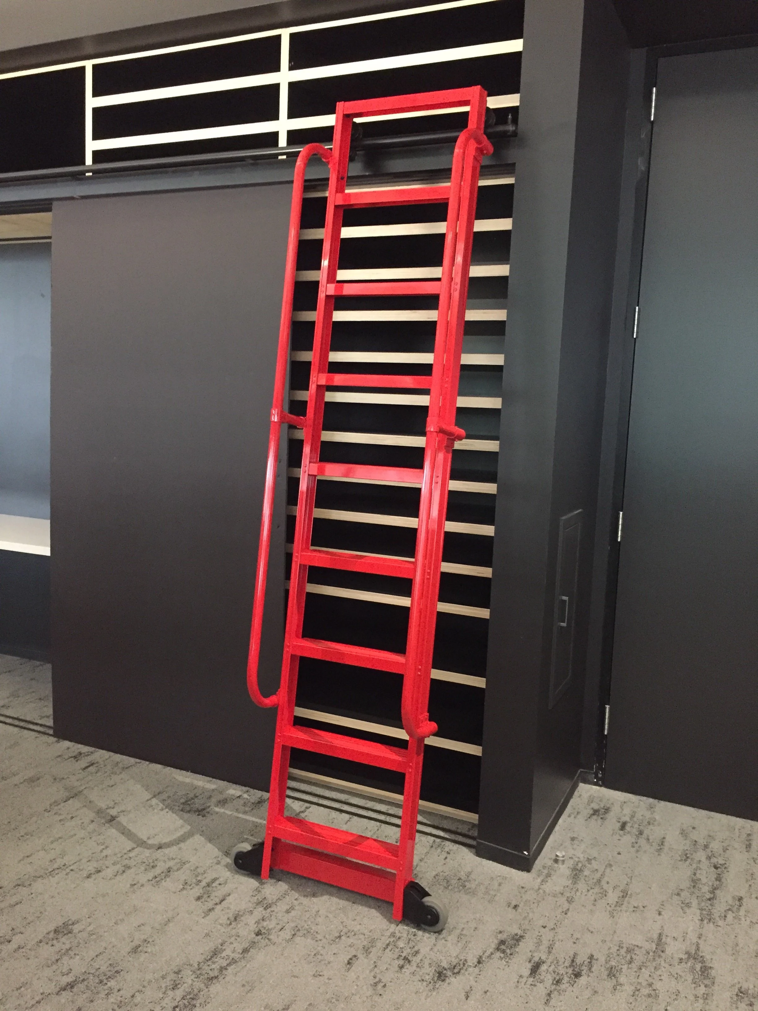 A red rolling ladder leaning against a black wall with horizontal light wood slats, in an indoor space with a gray door to the right and a patterned gray carpet floor.