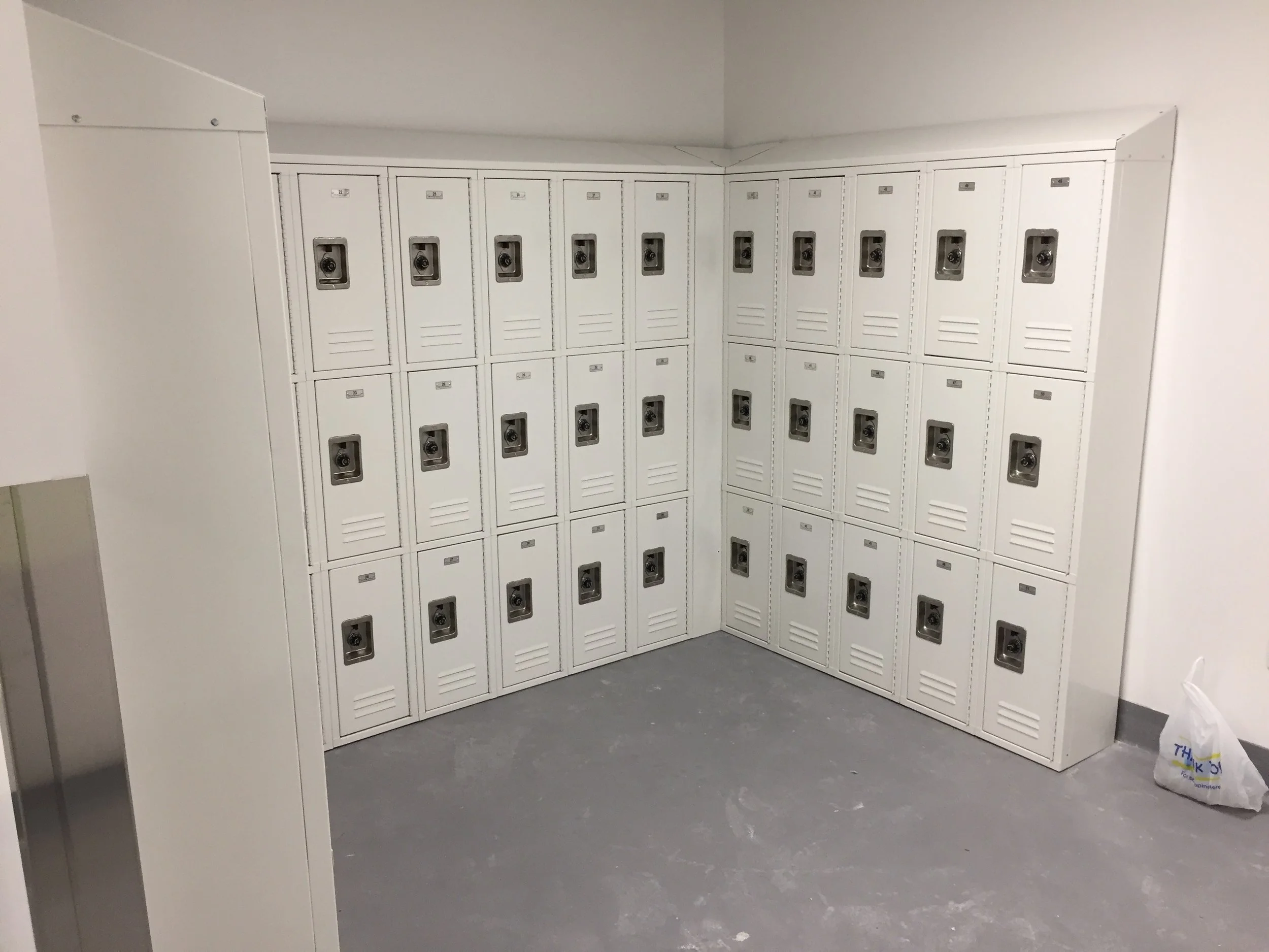 A corner of a room with white lockers arranged in a U-shape, each locker has a keyhole and a small label, with a gray floor and plain white walls, and a plastic bag on the floor.