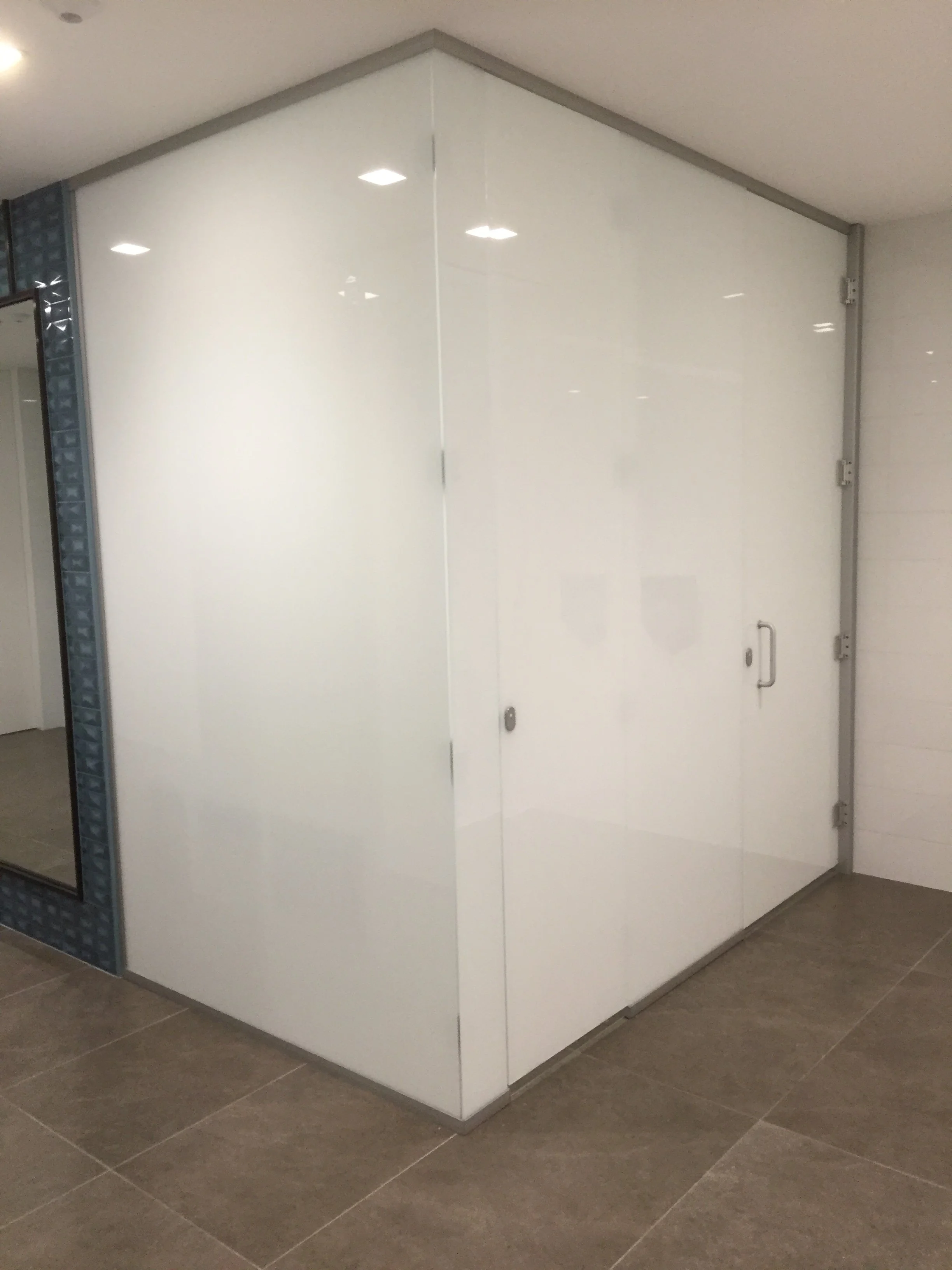 A modern interior space with a white, glossy wall that appears to be a large door or partition. The wall includes a handle and hinges, and the room features brown tiled flooring. The corner of the room is visible, with part of a glass block wall on t