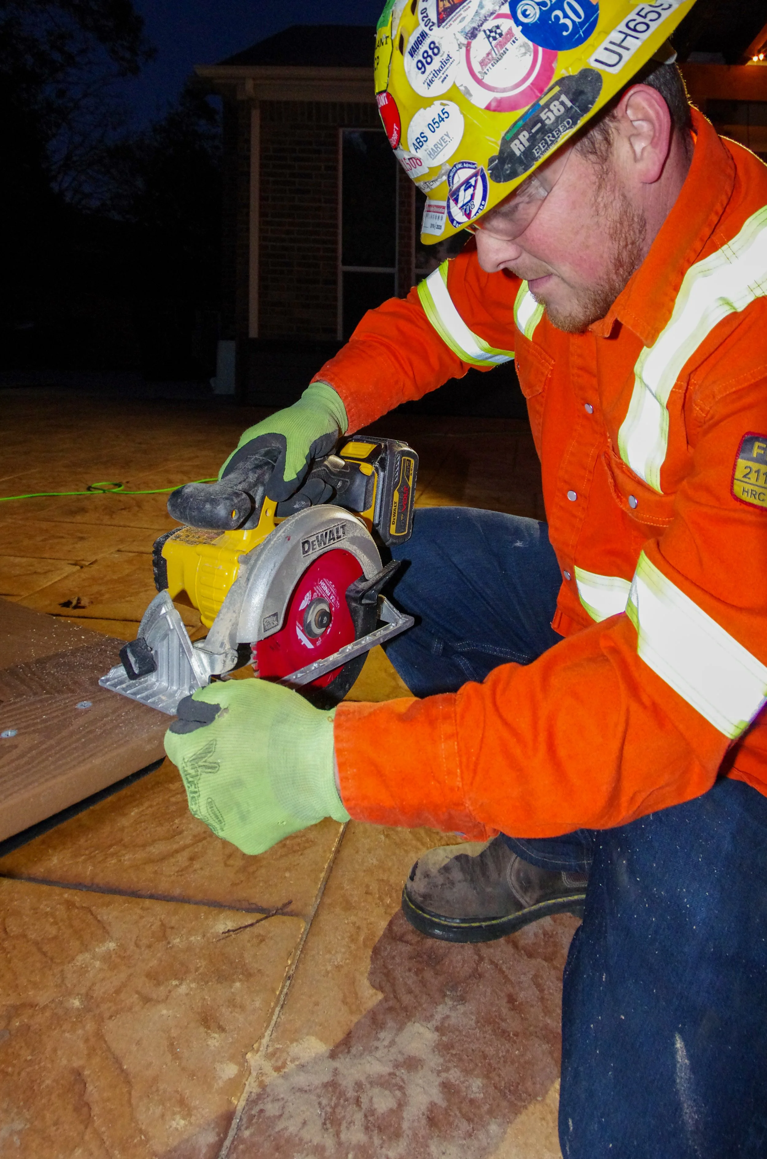A firefighter in an orange uniform and helmet with stickers is kneeling on a tiled floor and cutting wood with a circular saw. It is night time outside a house.