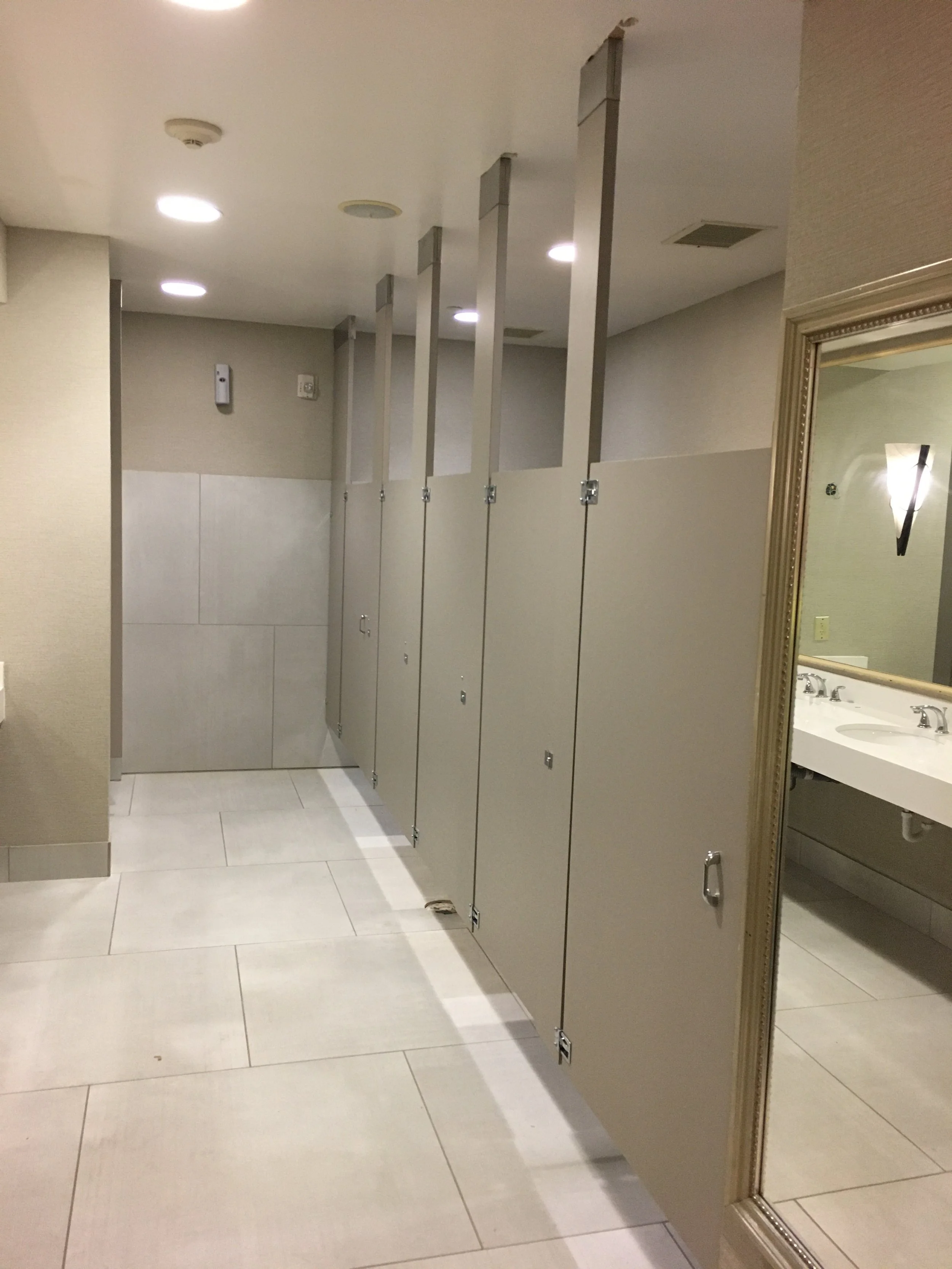 Public restroom with beige walls, beige tiled floor, beige bathroom stall doors, and a large mirror with gold frame reflecting a sink and faucet.