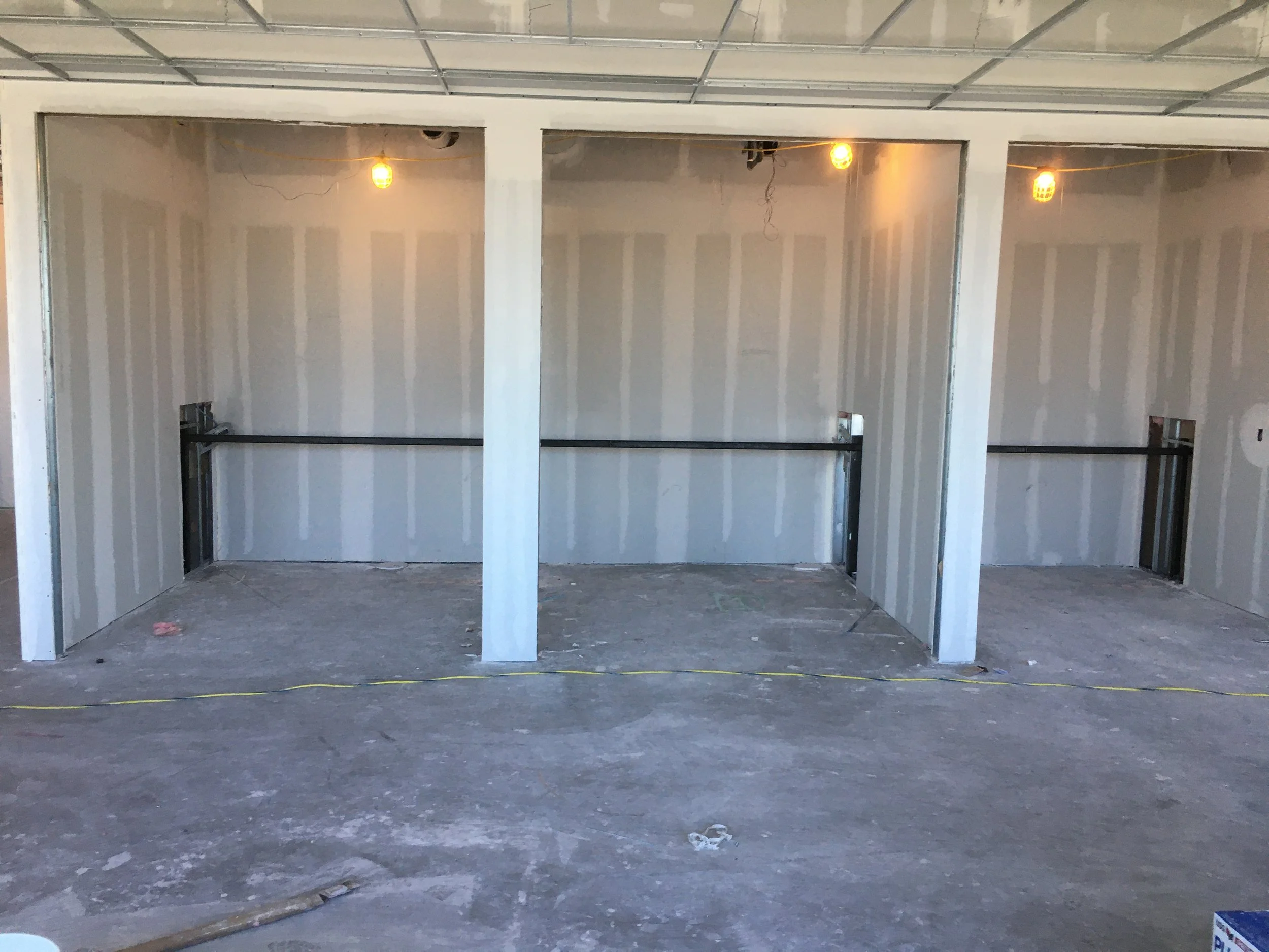 Interior of a building under construction with unfinished walls, a ceiling with visible framing, and hanging light bulbs. There are support beams and electrical outlets but no furnishings or decor.