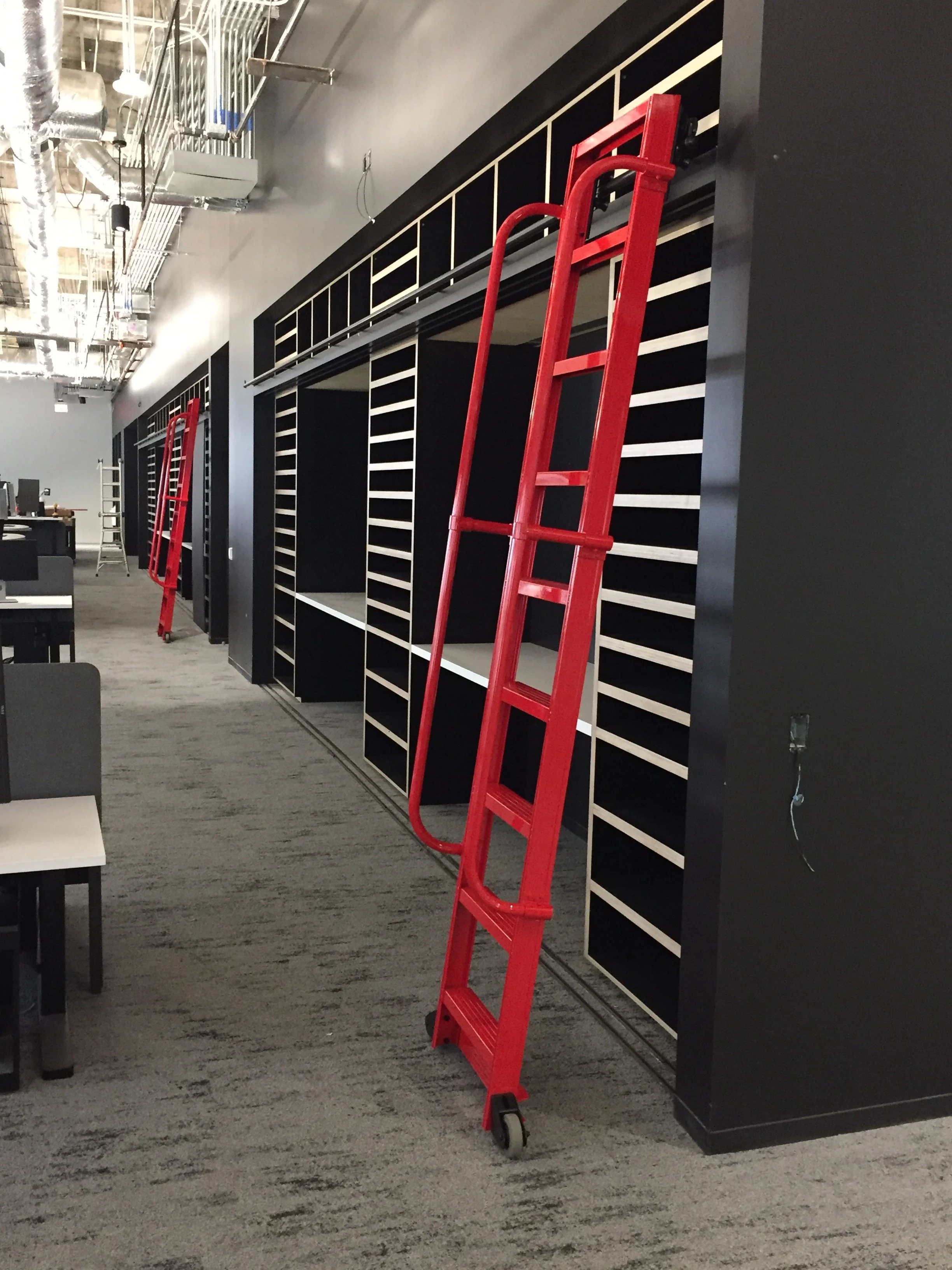 Red platform cart leaning against a black wall with shelves inside an office or retail space with exposed ceiling pipes and gray carpet.