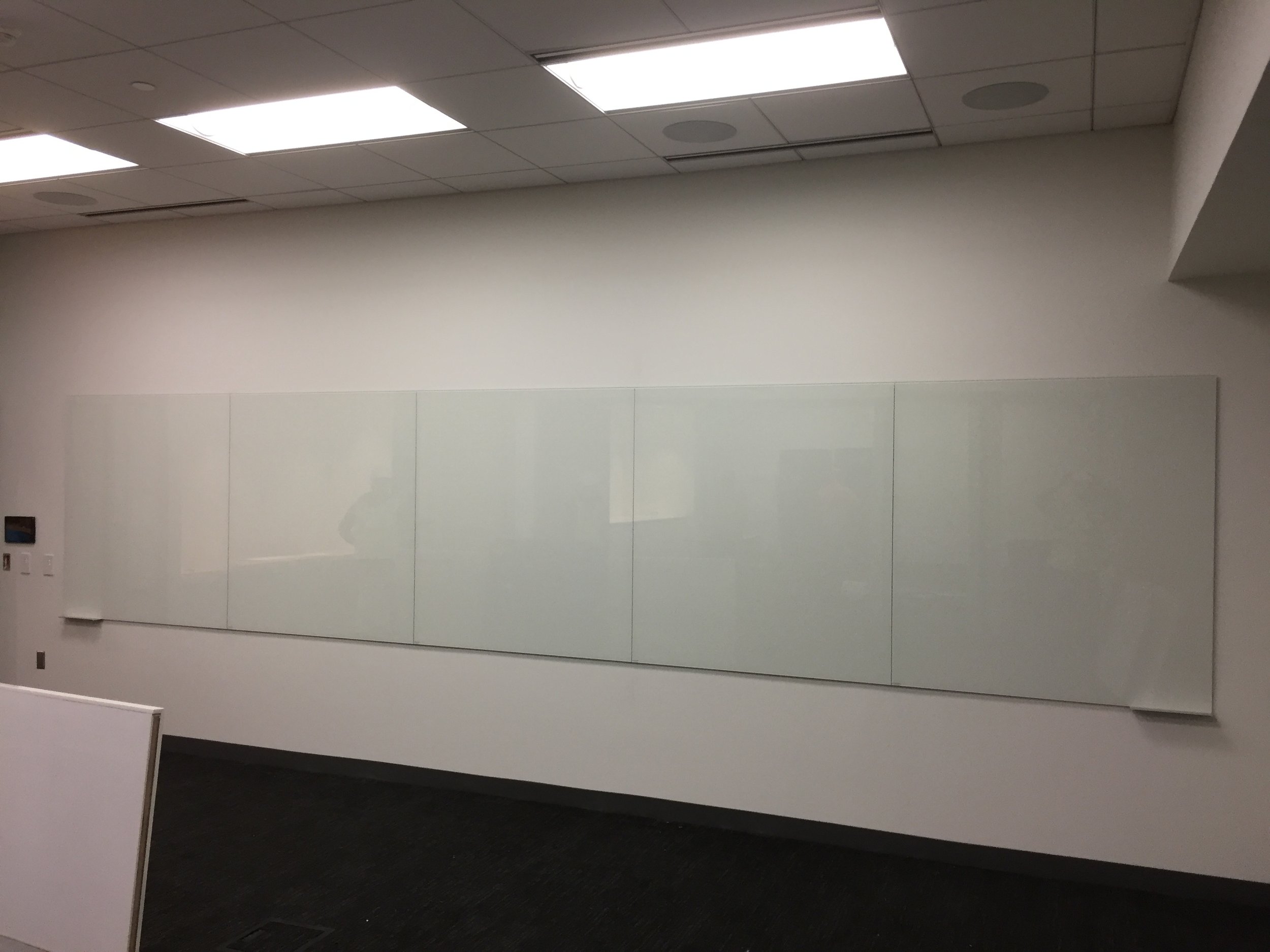 An empty conference room with a whiteboard or projection screen on the wall, ceiling lights, and a dark carpeted floor.