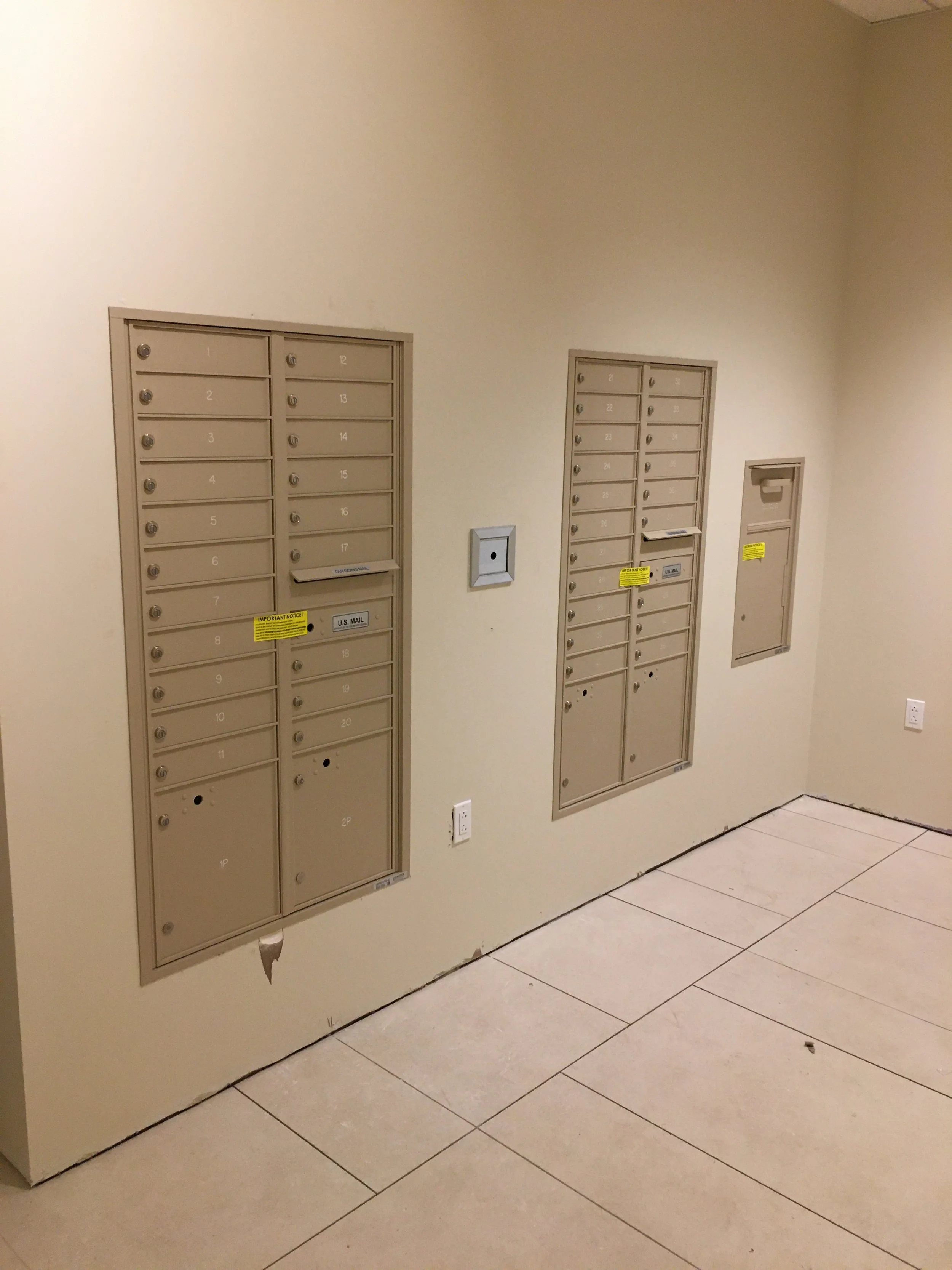 Wall-mounted mailboxes with numbered slots, a small mailbox door to the right, and an electrical outlet, all in a beige relay room with tiled floor.