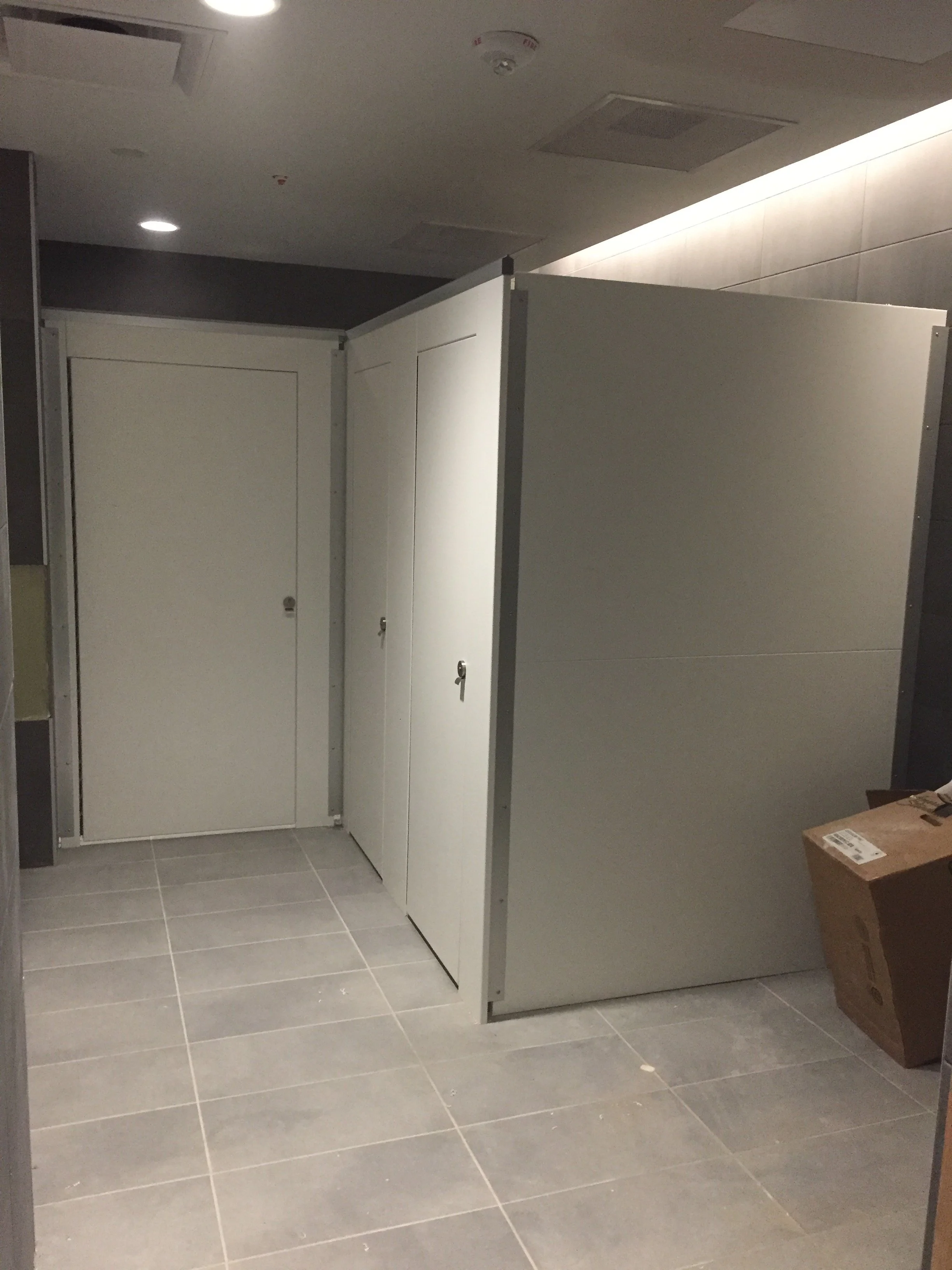 Public restroom with multiple white stalls, some with doors and some without. The floor is tiled with gray tiles, and there is a cardboard box on the right side. The ceiling has vents and a smoke detector.