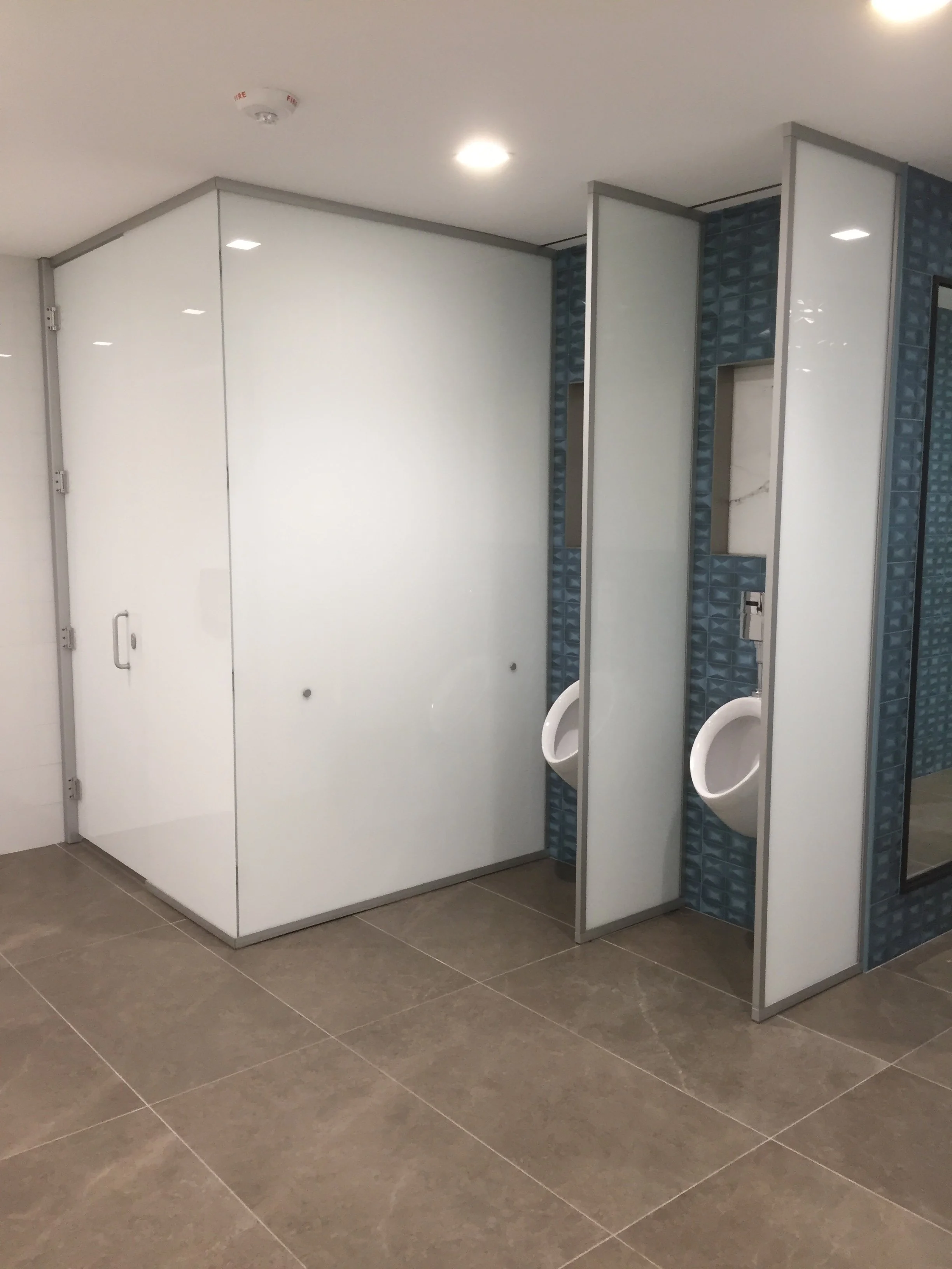 Public men's restroom with two urinals, white partition doors, blue tiled walls, and gray floor tiles.