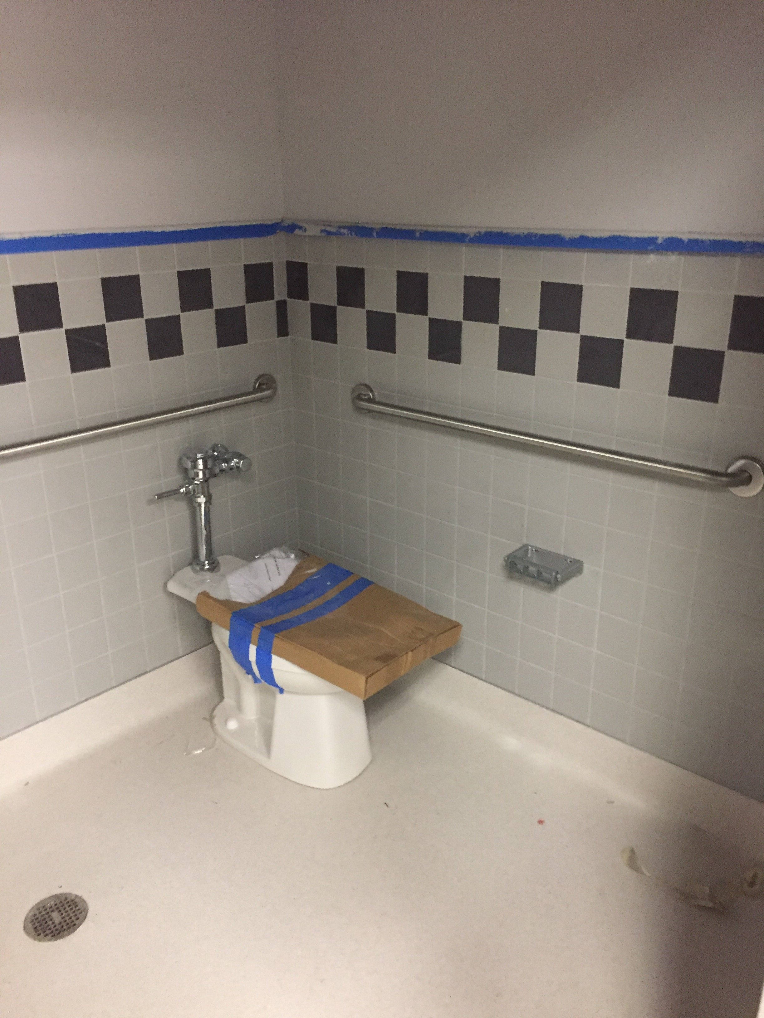 Bathroom under renovation with a toilet covered by painter's tape and a wooden plank, two metal grab bars attached to the tiled wall, a small metal soap dish, and blue painter's tape along the wall edge.