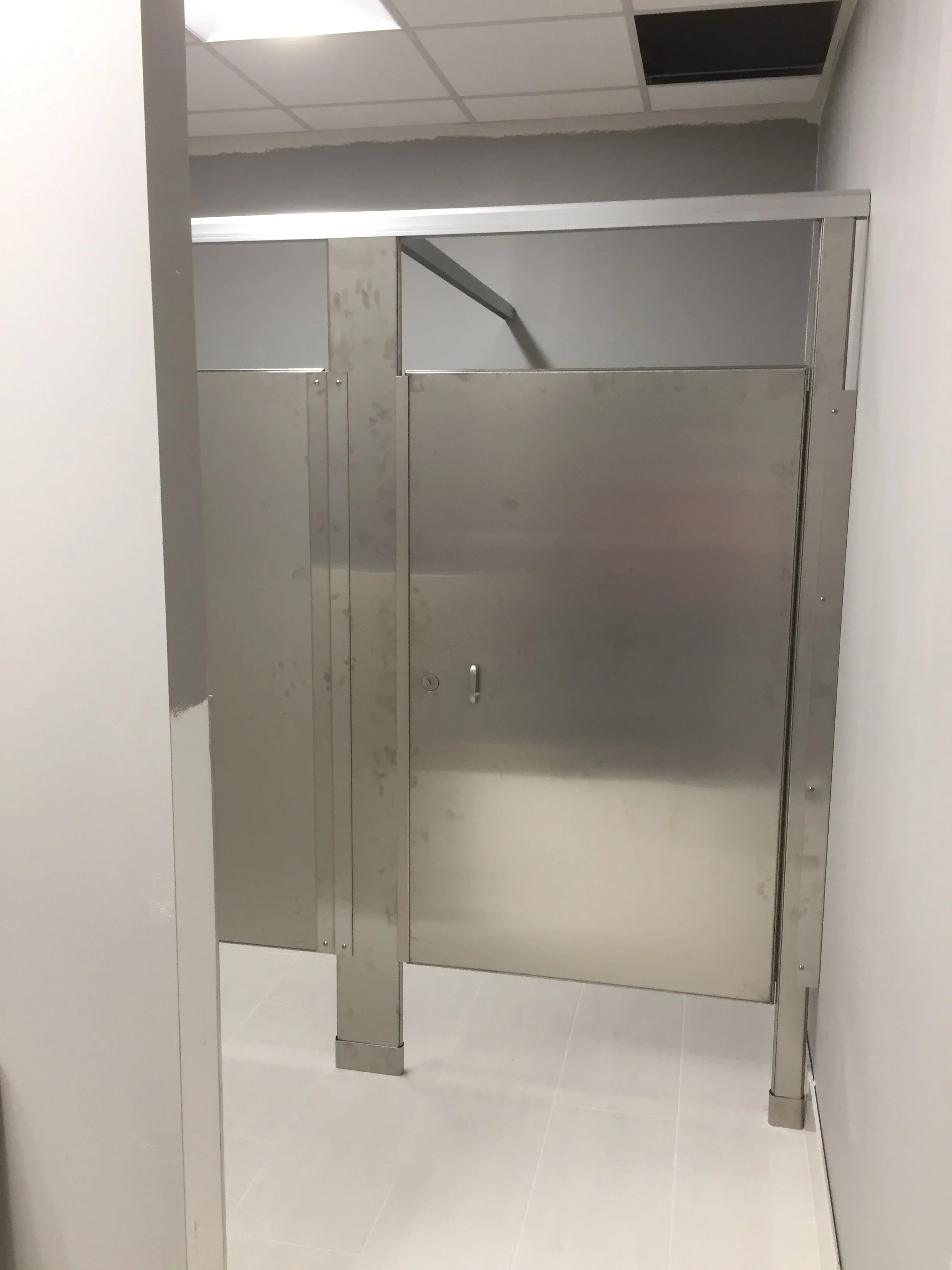 Stainless steel restroom stall divider and door, with a handle and lock, in a modern bathroom with tiled floor and false ceiling.