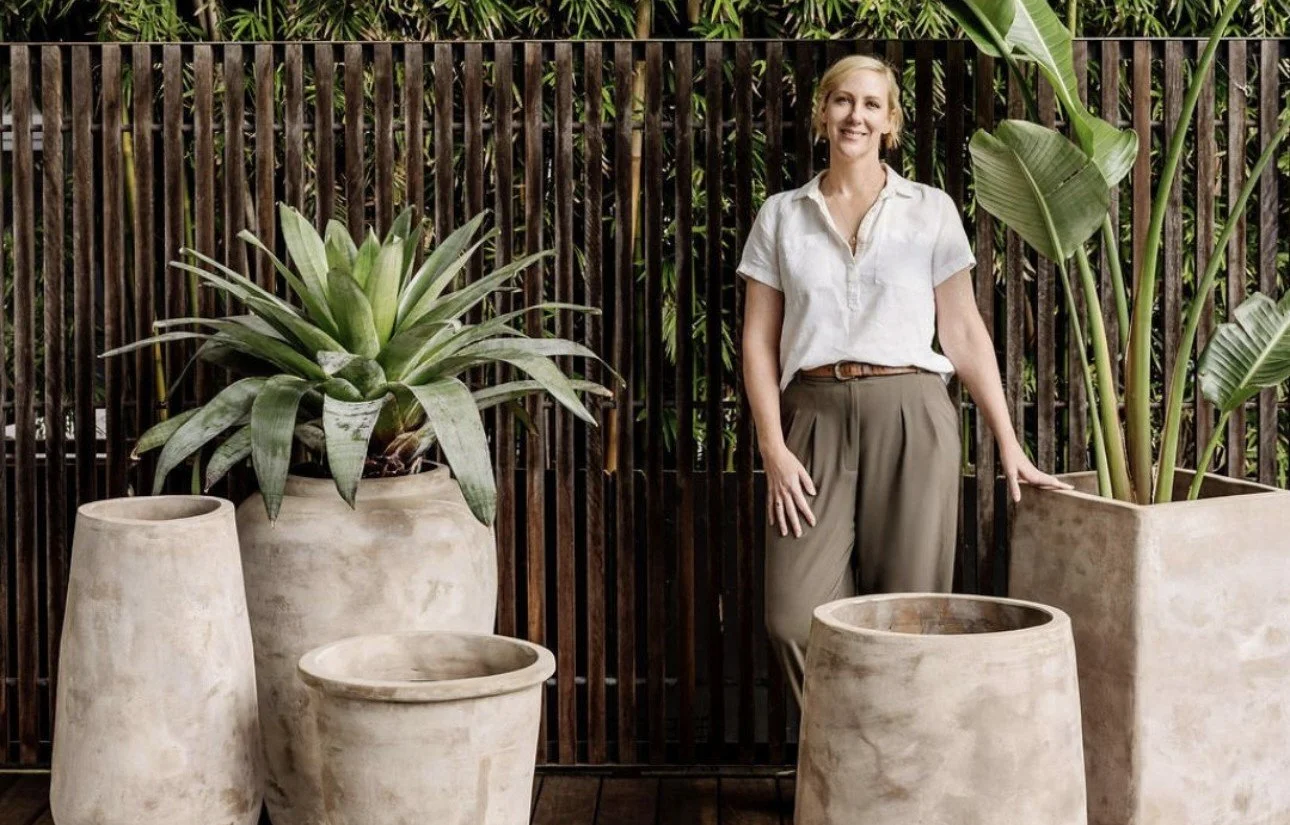 A woman standing next to large potted plants outside, with a wooden fence in the background.