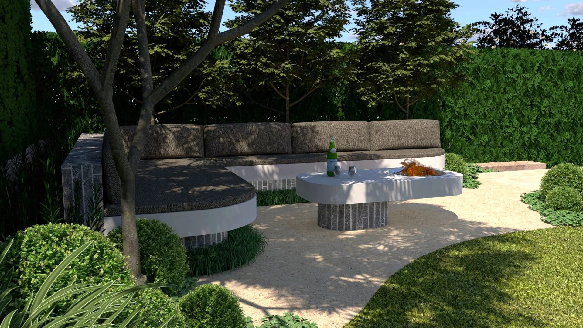 An outdoor patio scene featuring a curved sectional sofa with brown cushions, a white round coffee table with a brick base, surrounded by lush greenery, trees providing shade, a bottle and glasses on the table, and a gravel pathway.