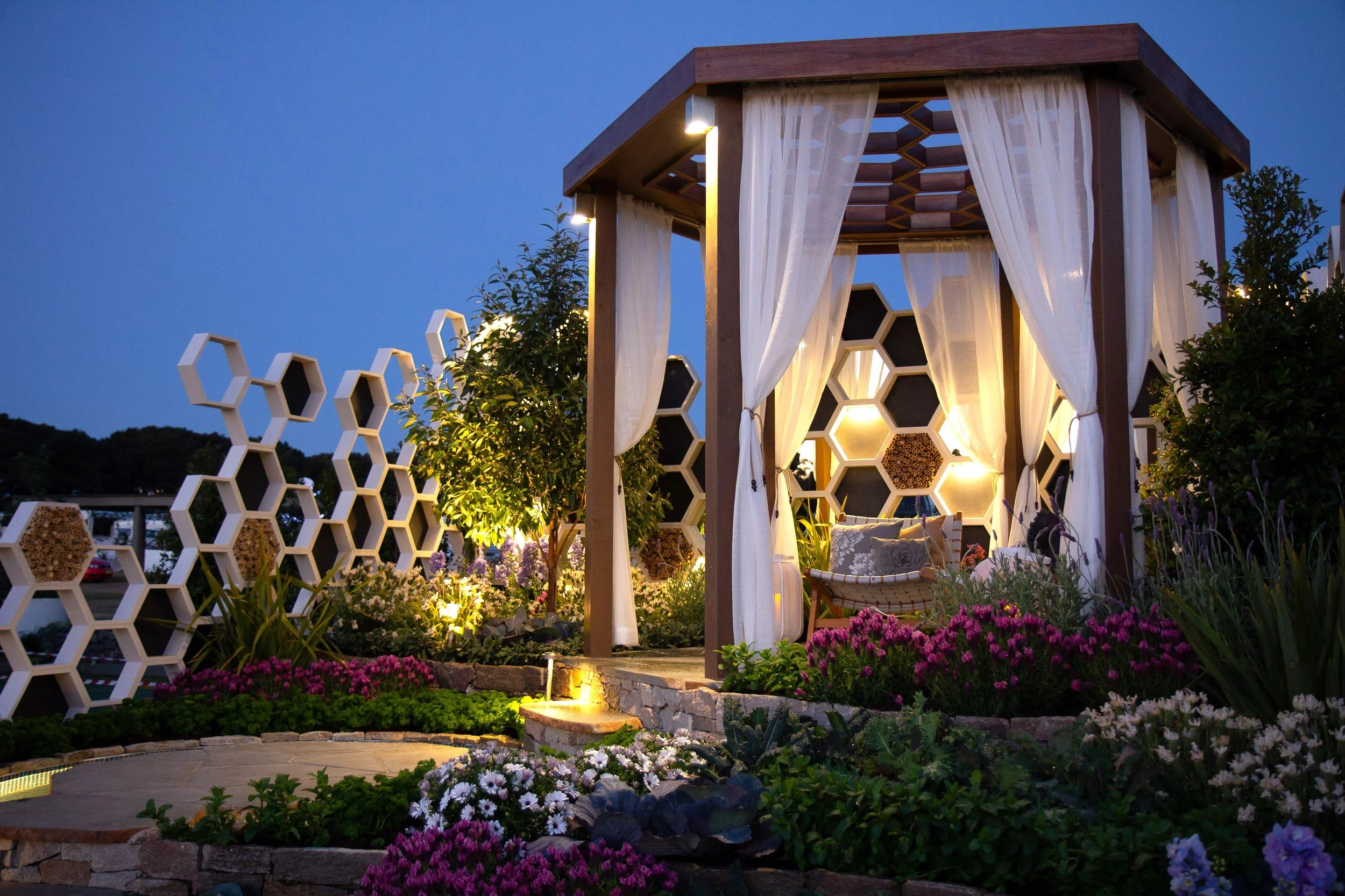 An outdoor seating area with a wooden gazebo with white curtains, surrounded by colorful flowers and decorative honeycomb-patterned walls, at dusk.
