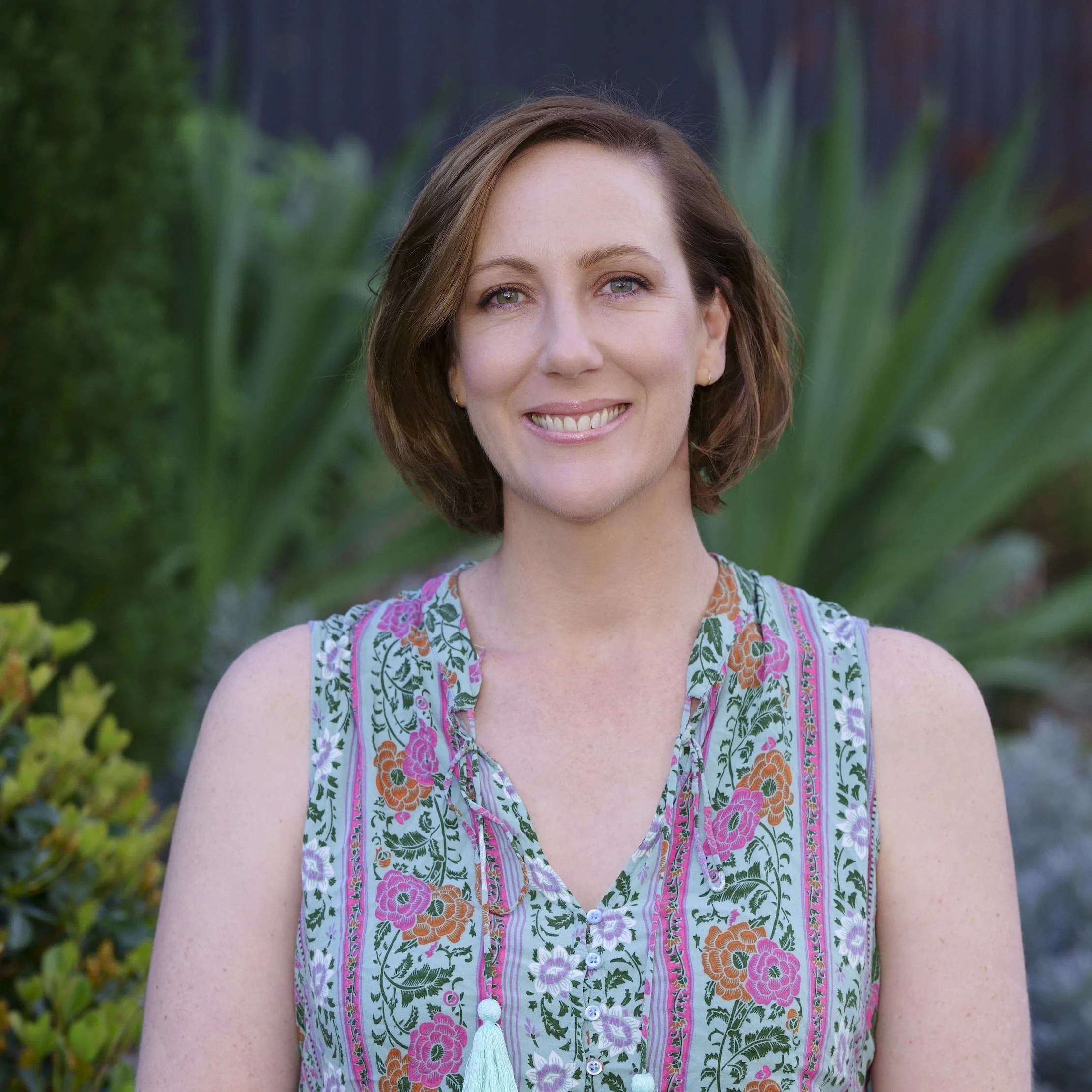 A woman with shoulder-length brown hair and blue eyes smiling outdoors. She is wearing a sleeveless floral top with pink, purple, orange, and green patterns and has a light blue tassel necklace. The background features green foliage.