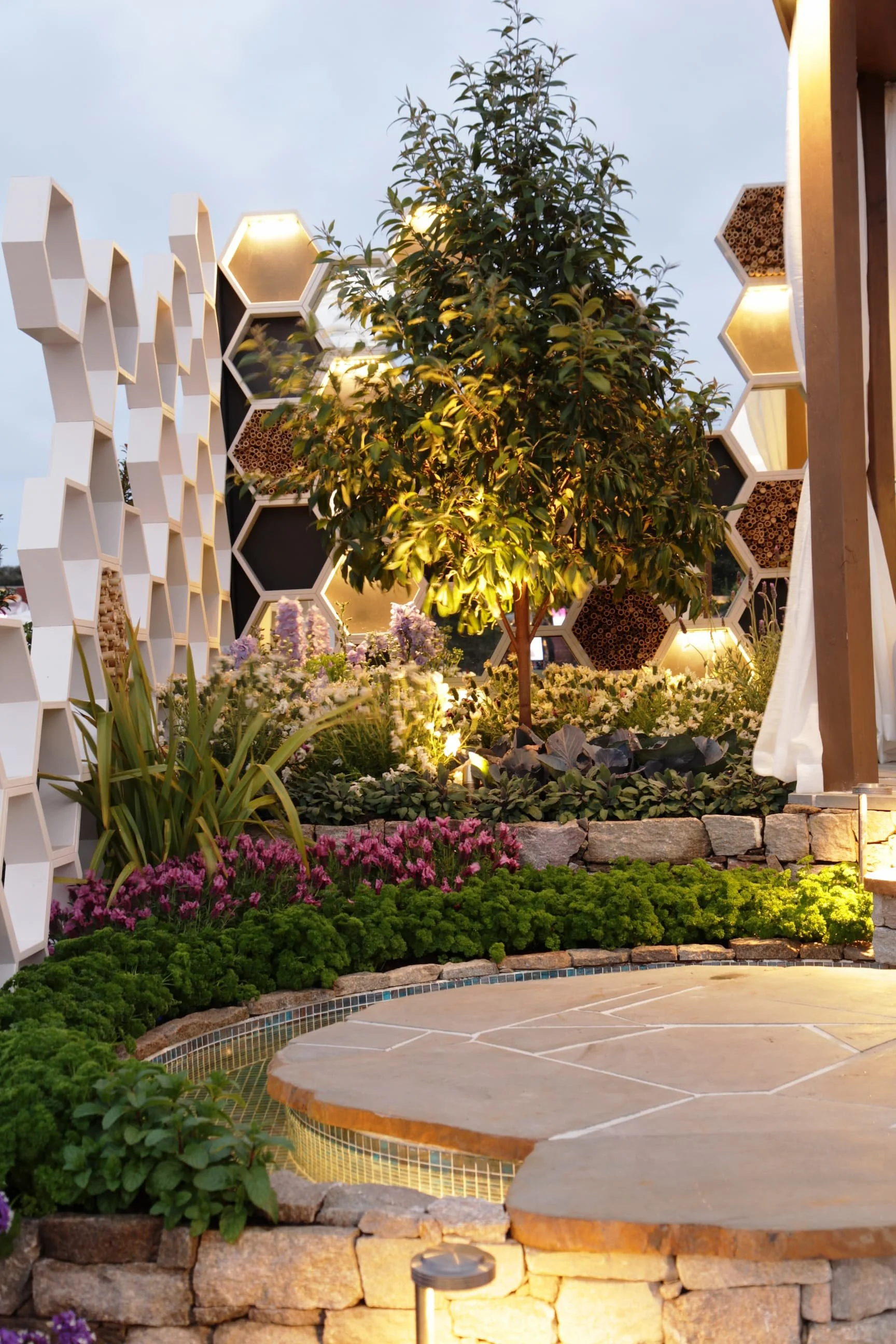 Nighttime outdoor garden scene with a tree, illuminated plants, flowers, and decorative geometric structures with lights.
