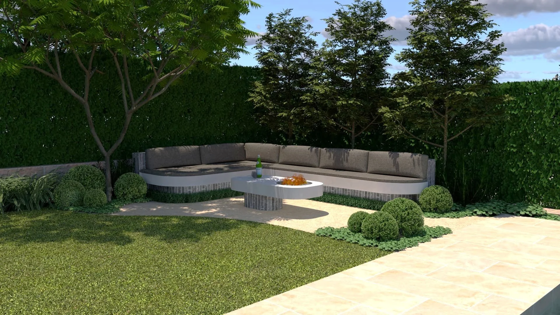 A backyard patio with a semicircular gray outdoor sofa with brown cushions, a white round coffee table with a bottle and orange decoration, surrounded by green grass, bushes, and trees, with a blue sky and some clouds overhead.