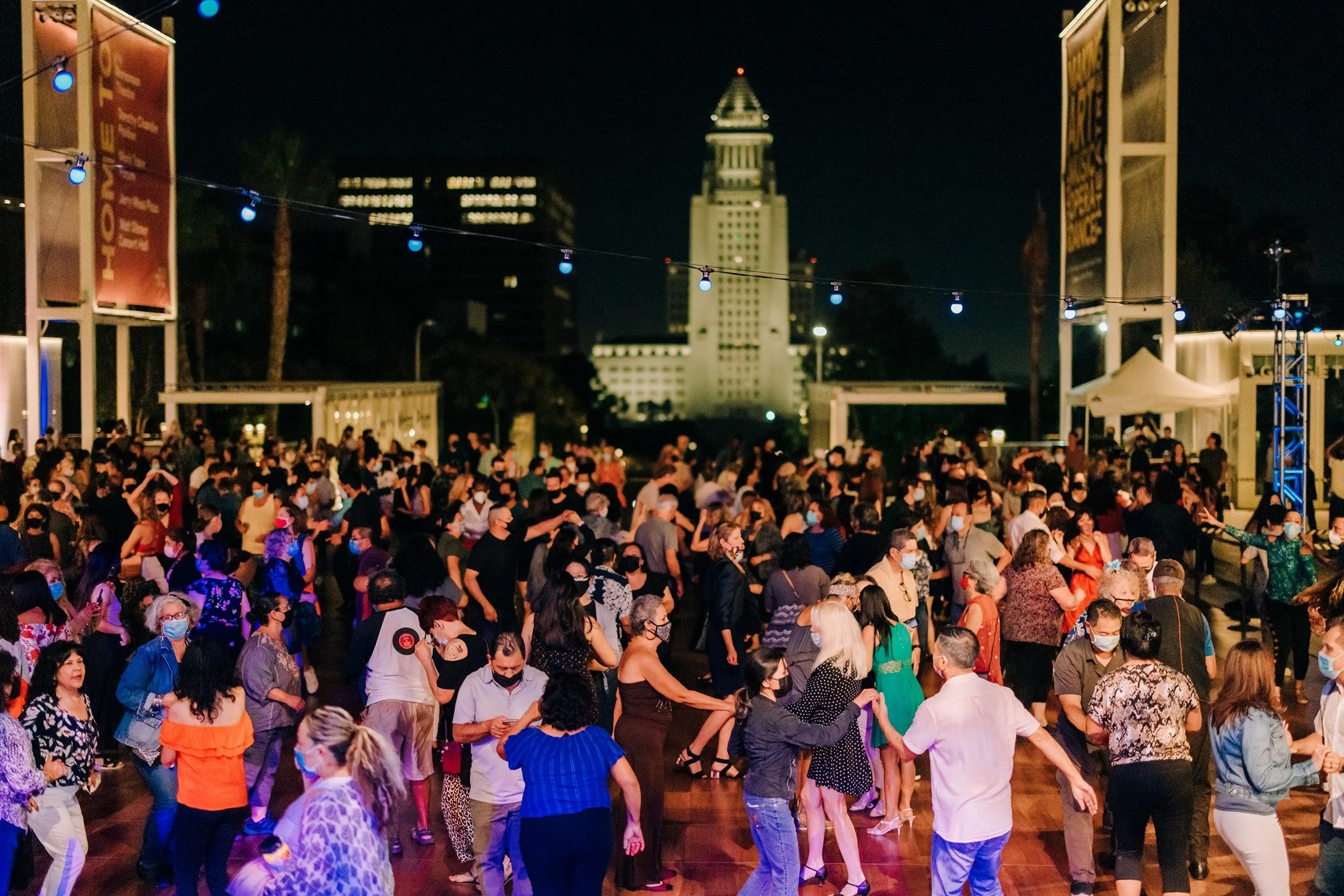 Groups of people dancing salsa on a wooden dance floor with a view of Los Angeles City Hall in the background