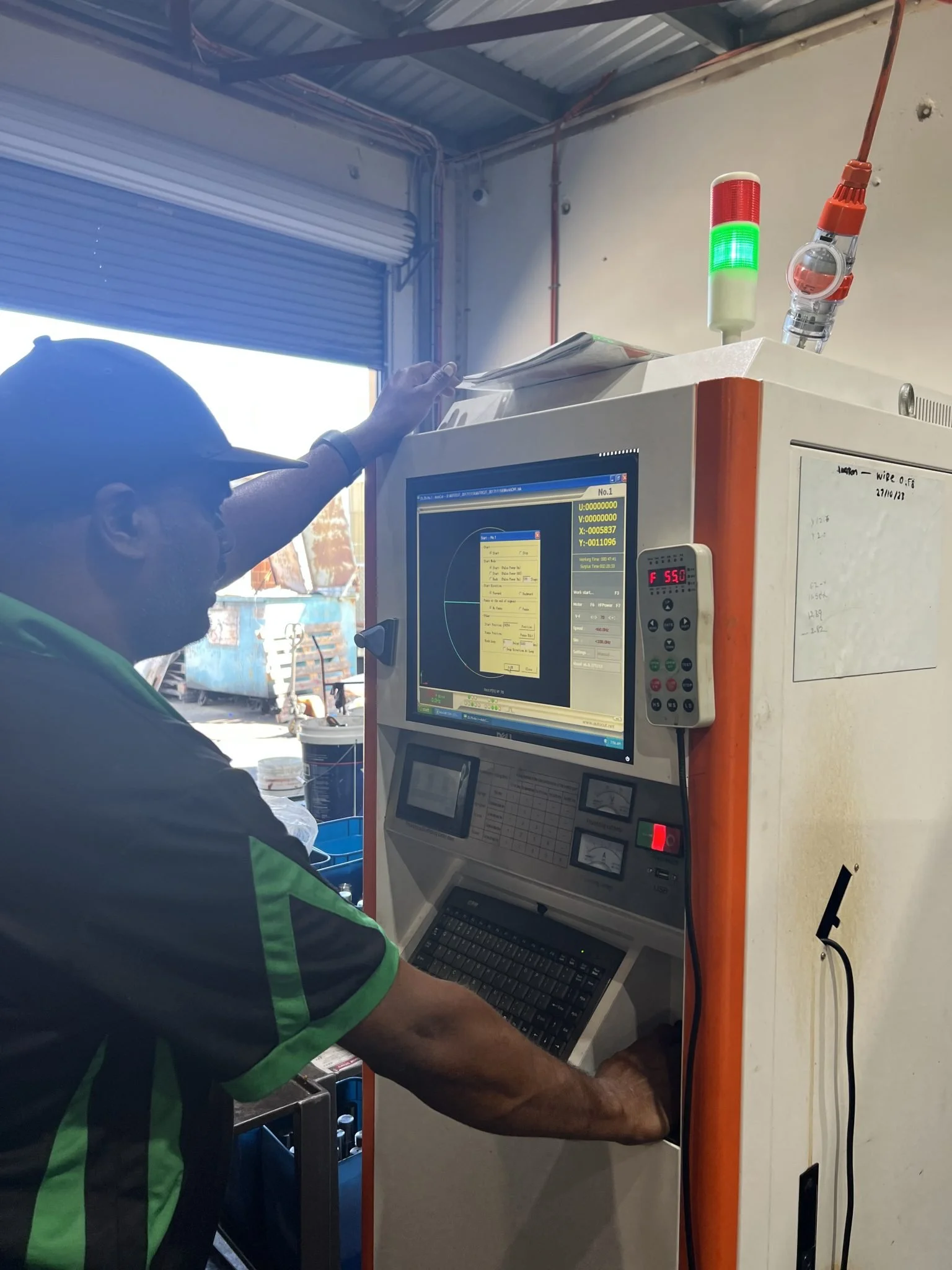 A person wearing a dark shirt with green stripes is working on a computer with a monitor displaying data and graphs. The workspace has a white machine with a digital display showing 'F 55.0' and a multi-colored light indicator with red and green lights. In the background, there is a partially open garage door, a large blue container, and some construction or industrial equipment outside.