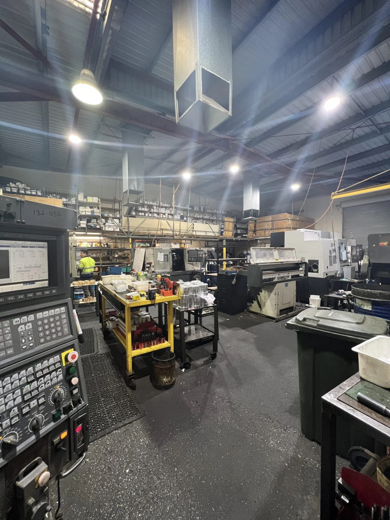 Industrial workshop or factory with machinery, workbenches, and equipment. Bright overhead lights illuminate the space. A person wearing a safety vest is working at a station.