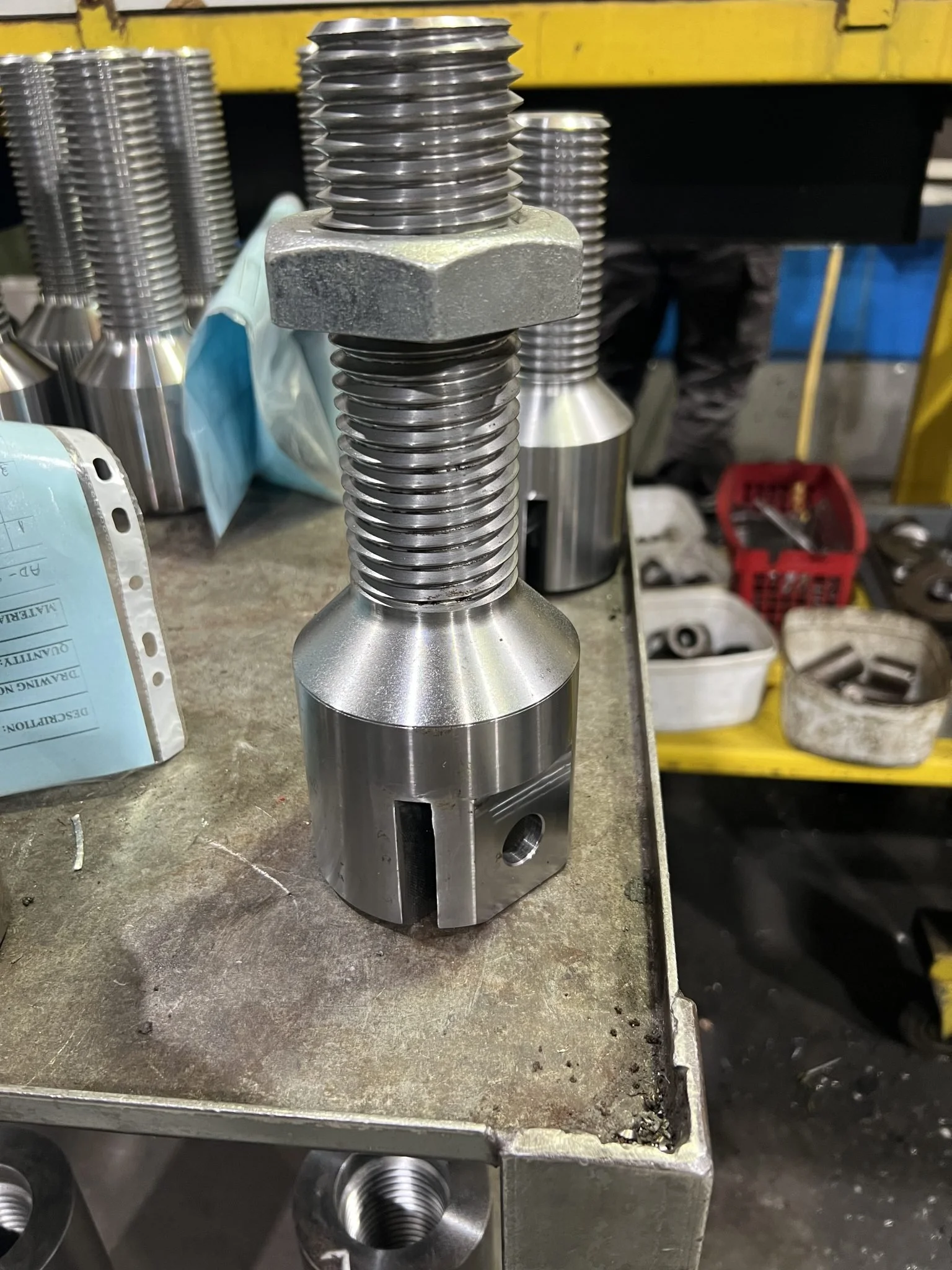 Close-up of a large machined metal bolt with a textured hexagonal head and a threaded shaft, resting on a workbench in a workshop.