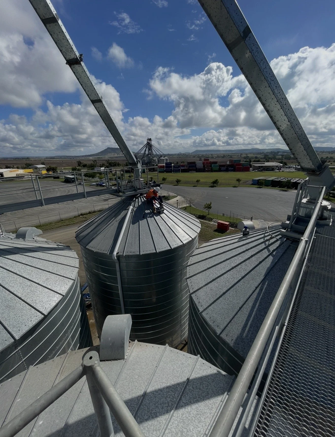 A person working on the roof of a large industrial building with metal tanks and structures, under a partly cloudy sky.
