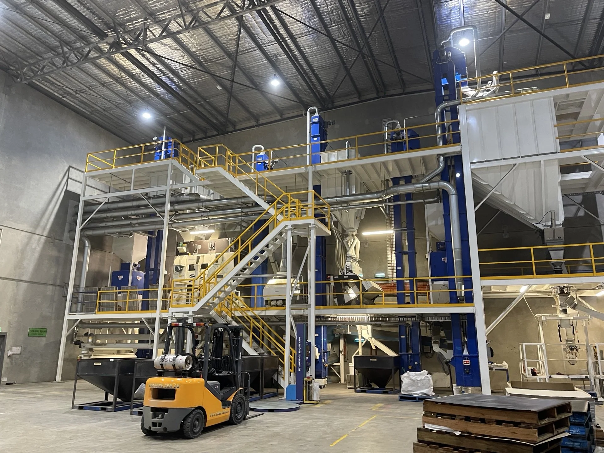 Industrial warehouse with metal stairs and platforms painted yellow, blue machinery, a forklift, and pallets on the concrete floor under a high ceiling.