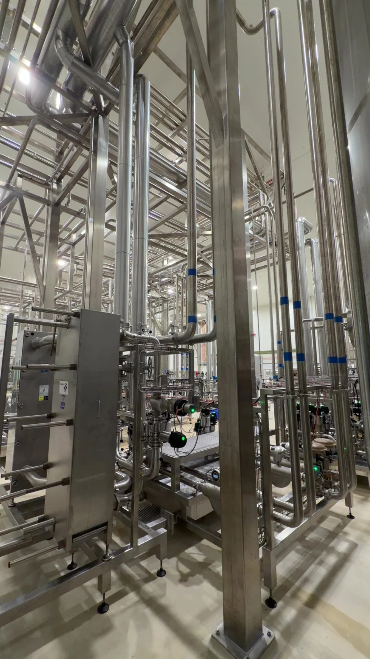 Industrial stainless steel piping system with interconnected pipes, valves, and gauges in a factory or processing plant.