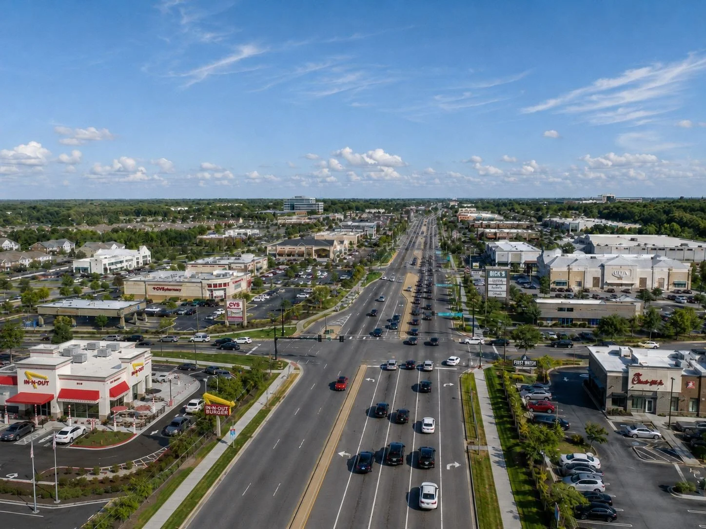 Aerial view of a busy commercial shopping center with multiple stores, parking lots, and cars on a main road under a partly cloudy sky.