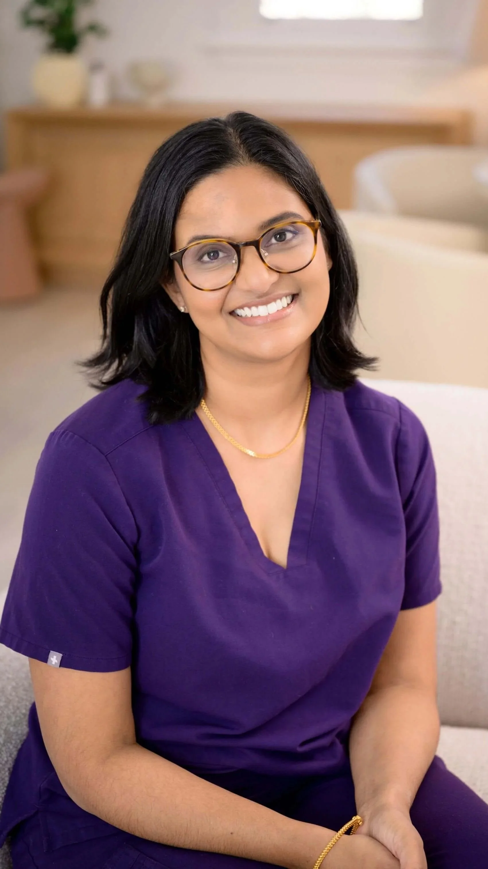 A woman with black hair, glasses, and a purple scrubs uniform sitting on a beige chair in a room with a wooden cabinet and a window in the background. She is smiling and looking at the camera.