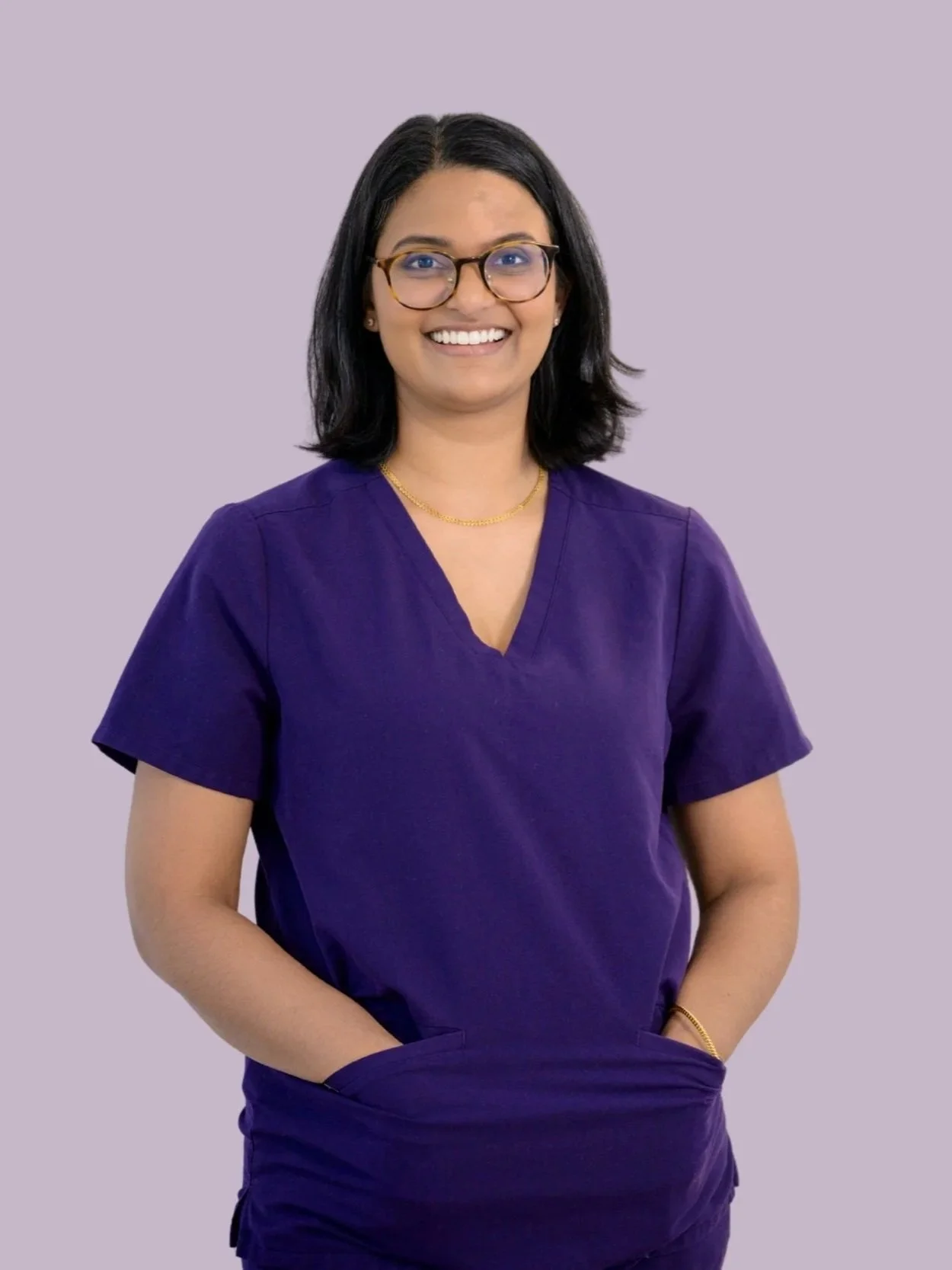 A woman in scrubs standing against a light purple background, smiling, with shoulder-length black hair, glasses, and a gold necklace.