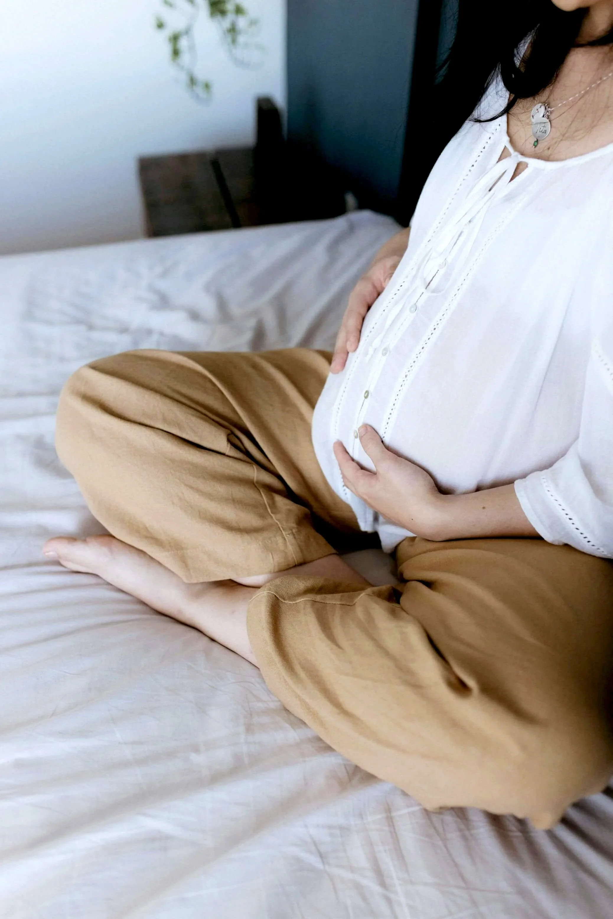 A person sitting cross-legged on a bed, holding their stomach with both hands, wearing a white blouse and tan pants, with necklaces, in a room with a bed and a bedside table.