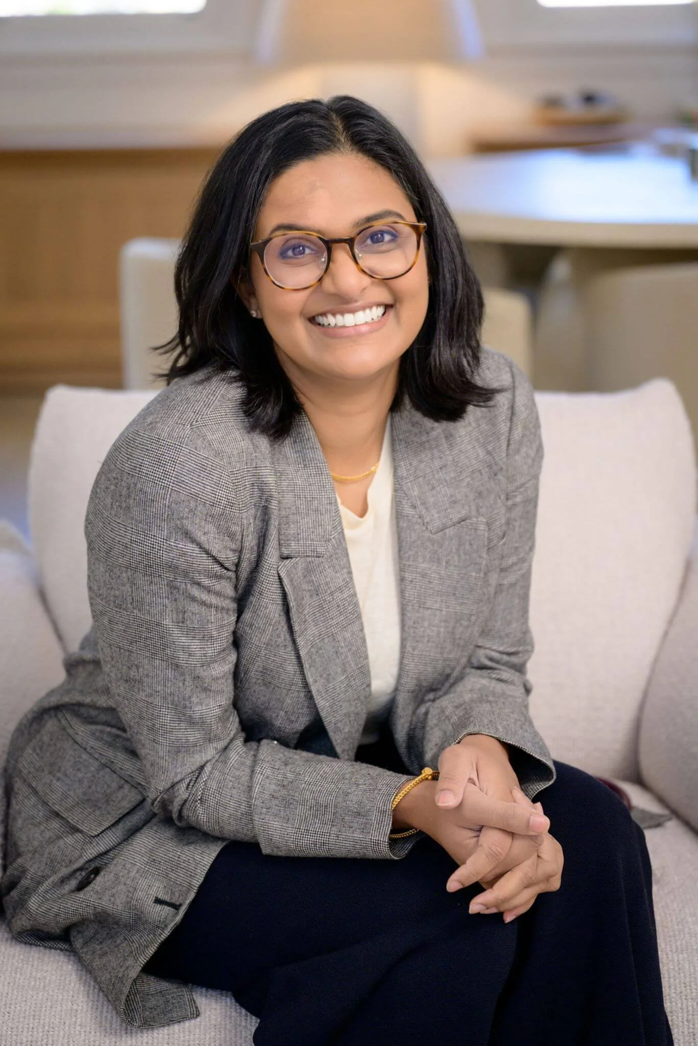 A woman with black hair, glasses, and a grey checkered blazer smiling, sitting on a beige couch in a well-lit room with wooden furniture in the background.