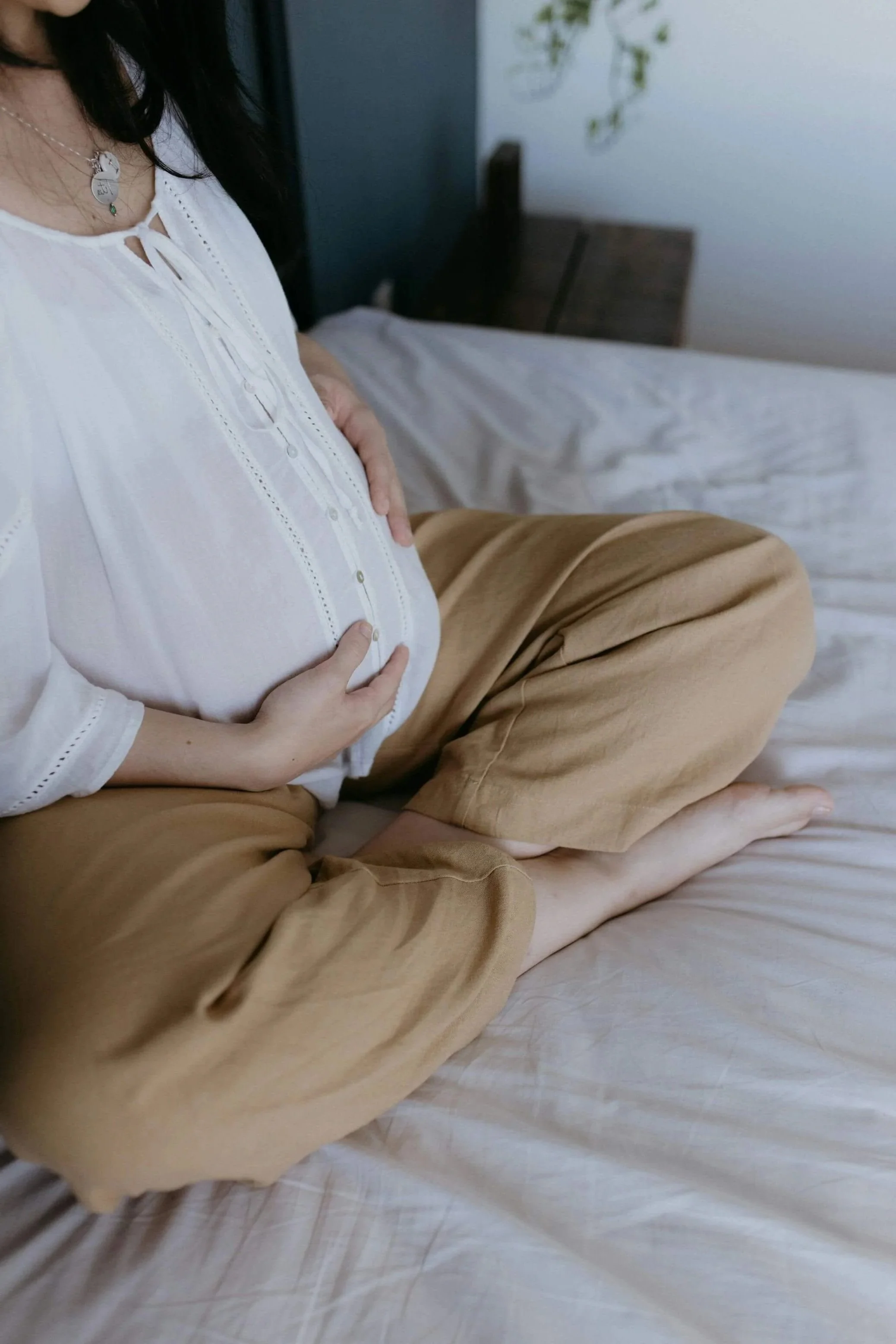 A pregnant woman sitting on a bed, touching her belly with both hands, wearing a white blouse and beige pants, with a pillow and a dark headboard in the background.