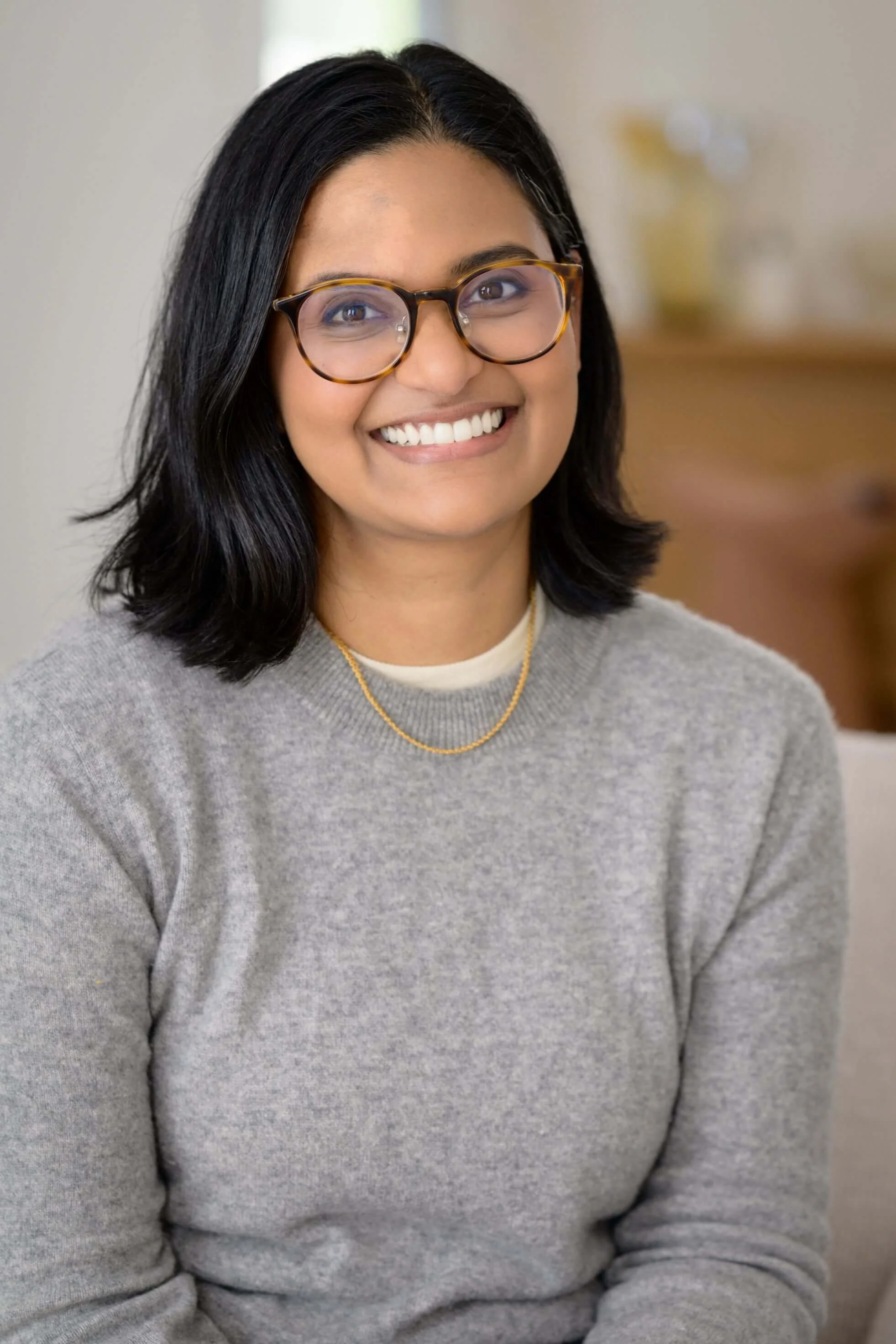 Close-up of a woman smiling, wearing round tortoiseshell eyeglasses, a gray sweater, a white shirt underneath, a gold necklace, and has shoulder-length black hair, in an indoor setting.