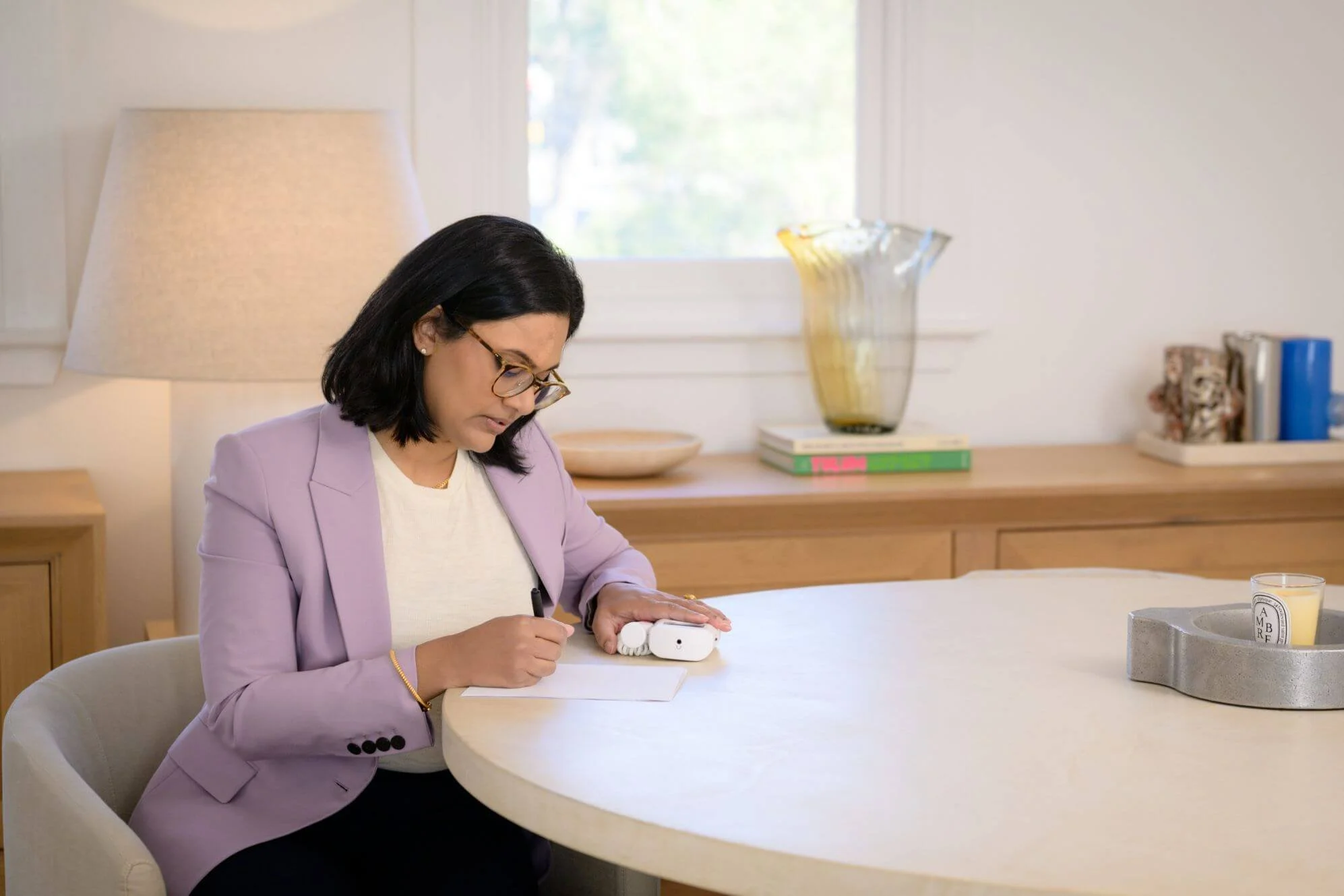 Woman with dark hair and glasses sits at a light-colored table, writing on a piece of paper, with a small device in her right hand. She is wearing a light purple blazer and a white top. The background includes a window, a large decorative glass vase, books, and a few small decorative items.
