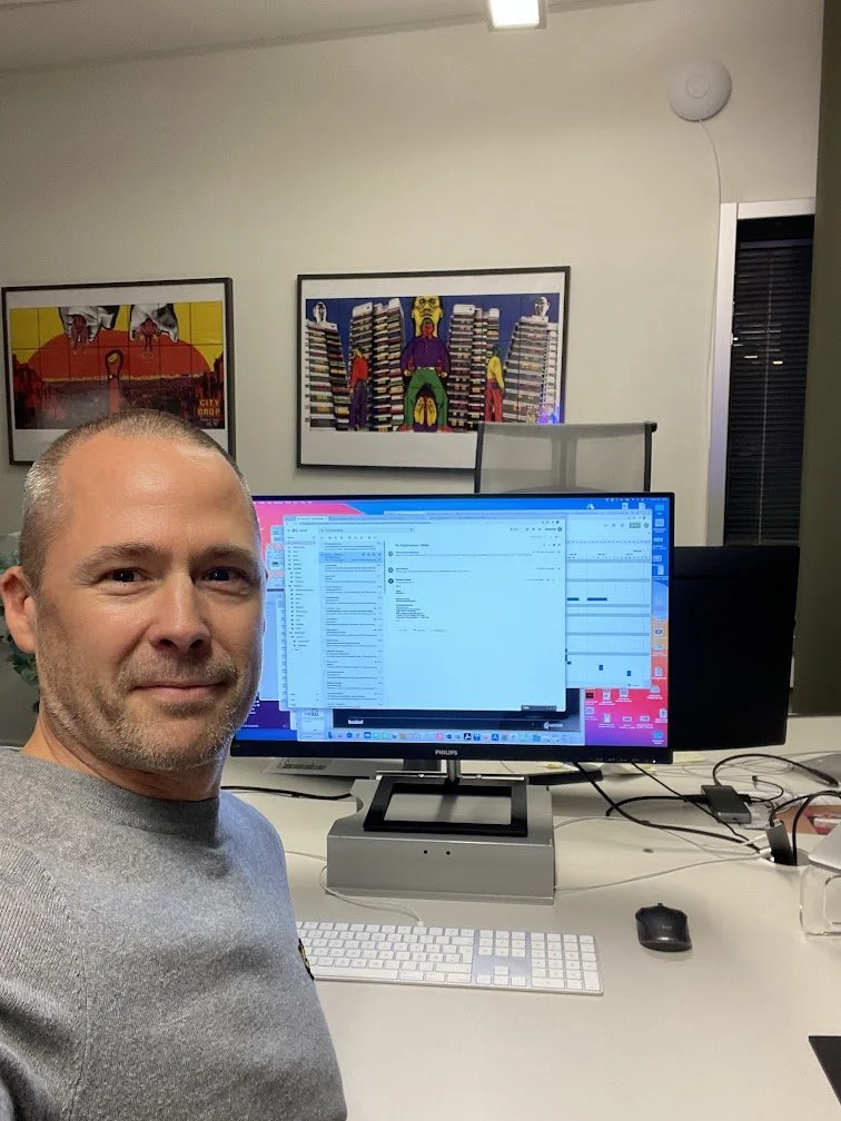 A man taking a selfie in an office with a computer monitor, keyboard, and mouse on the desk. There are colorful artworks hanging on the wall behind him.