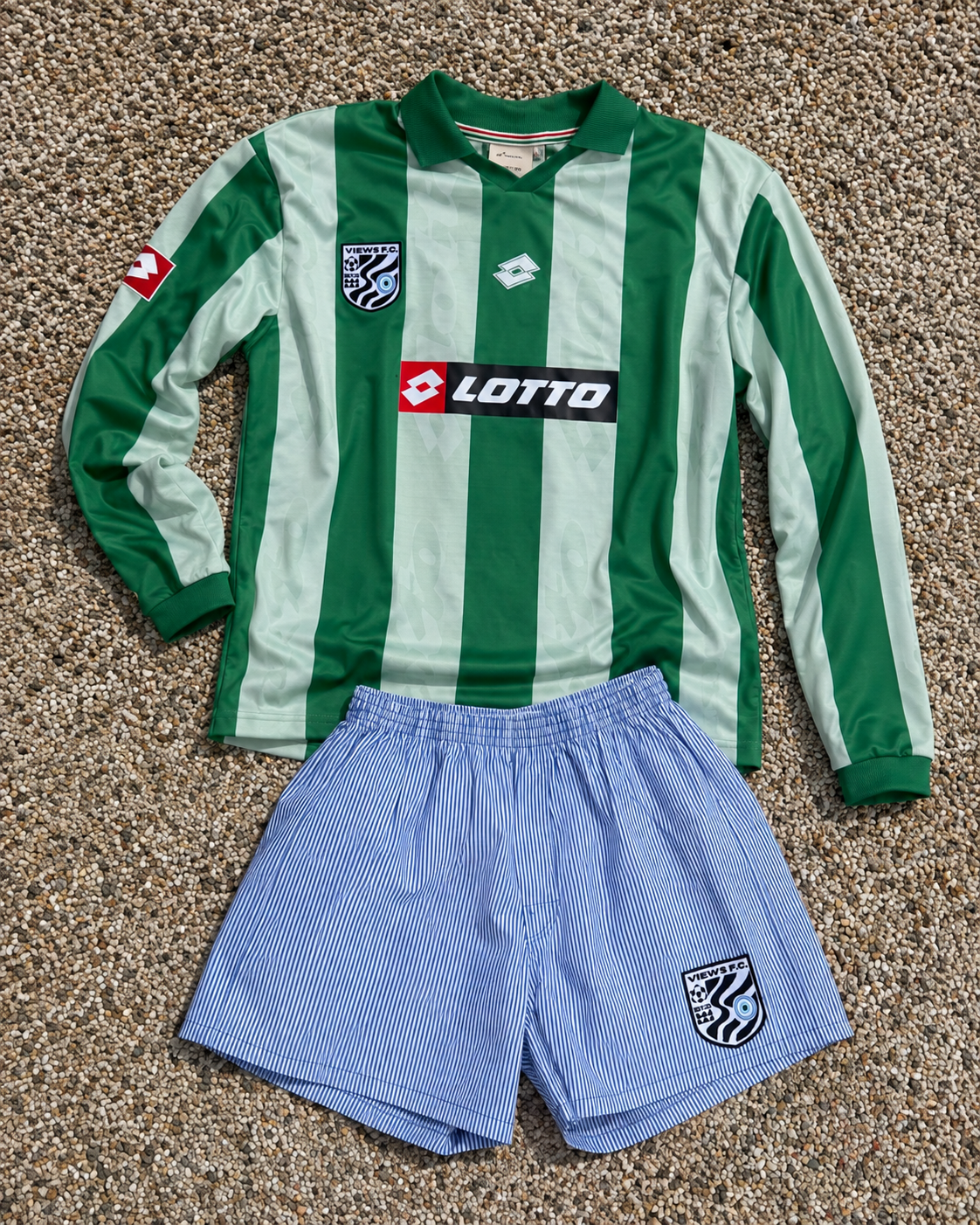 A green and white vertical striped soccer jersey with a collar, a black and white patch on the left chest, and sponsor logos, laid out on a gravel surface. A pair of blue and white striped shorts with a team crest on the left side are also shown bene