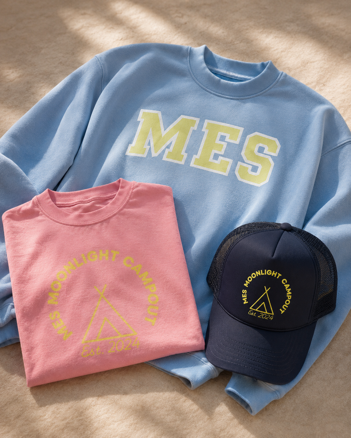 A blue sweatshirt with the word 'MES' in large letters, a pink T-shirt with 'MES Moonlight Campout Est. 2024' and a teepee graphic, and a navy baseball cap with the same text and graphic, all laid on a beige surface.