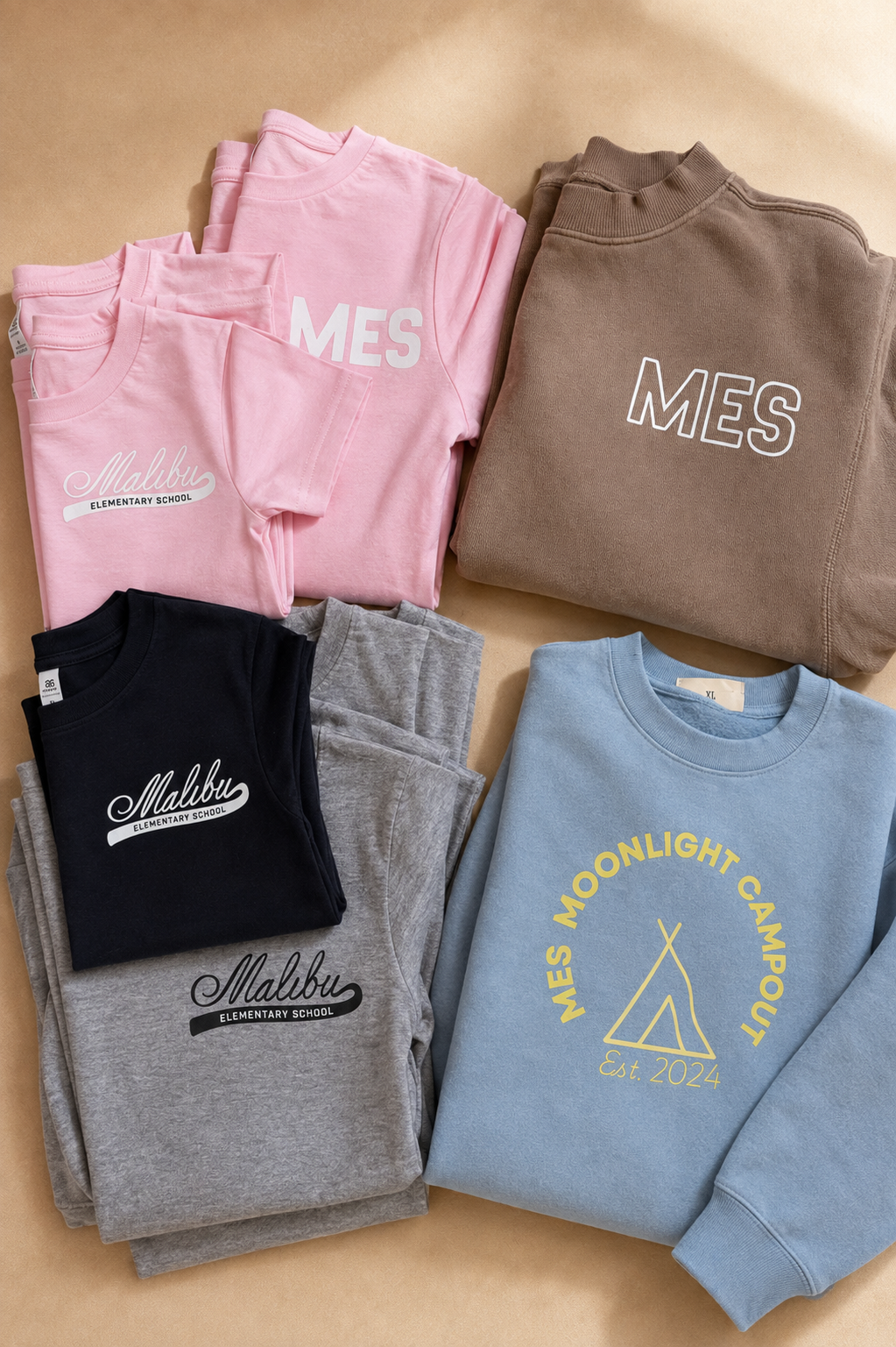 Stacked and folded colorful t-shirts and sweatshirts with school and camp logos on a beige background.