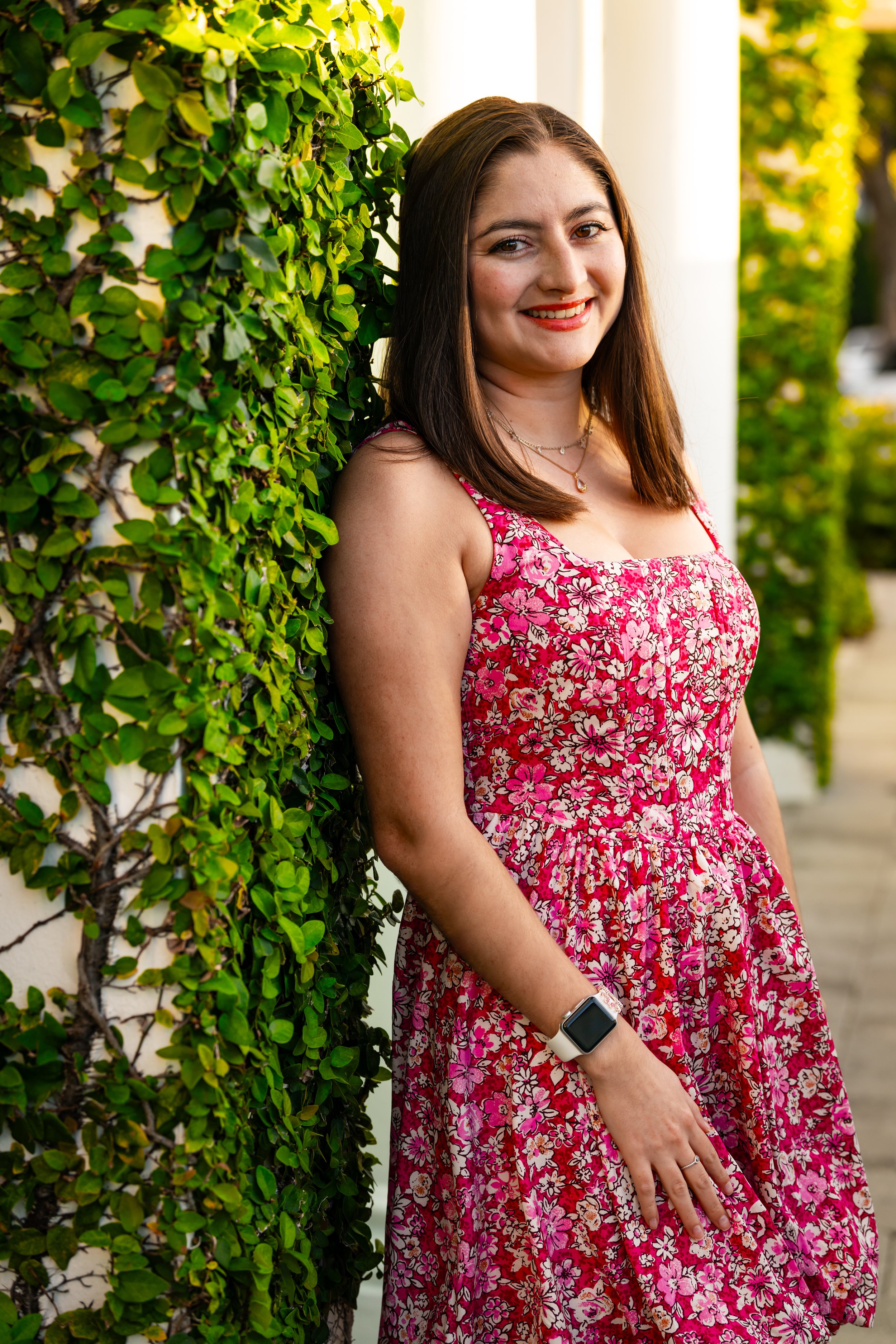 A young woman with shoulder-length brown hair smiling and leaning against a green hedge wall outdoors. She is wearing a pink floral dress, a necklace, and a smartwatch.