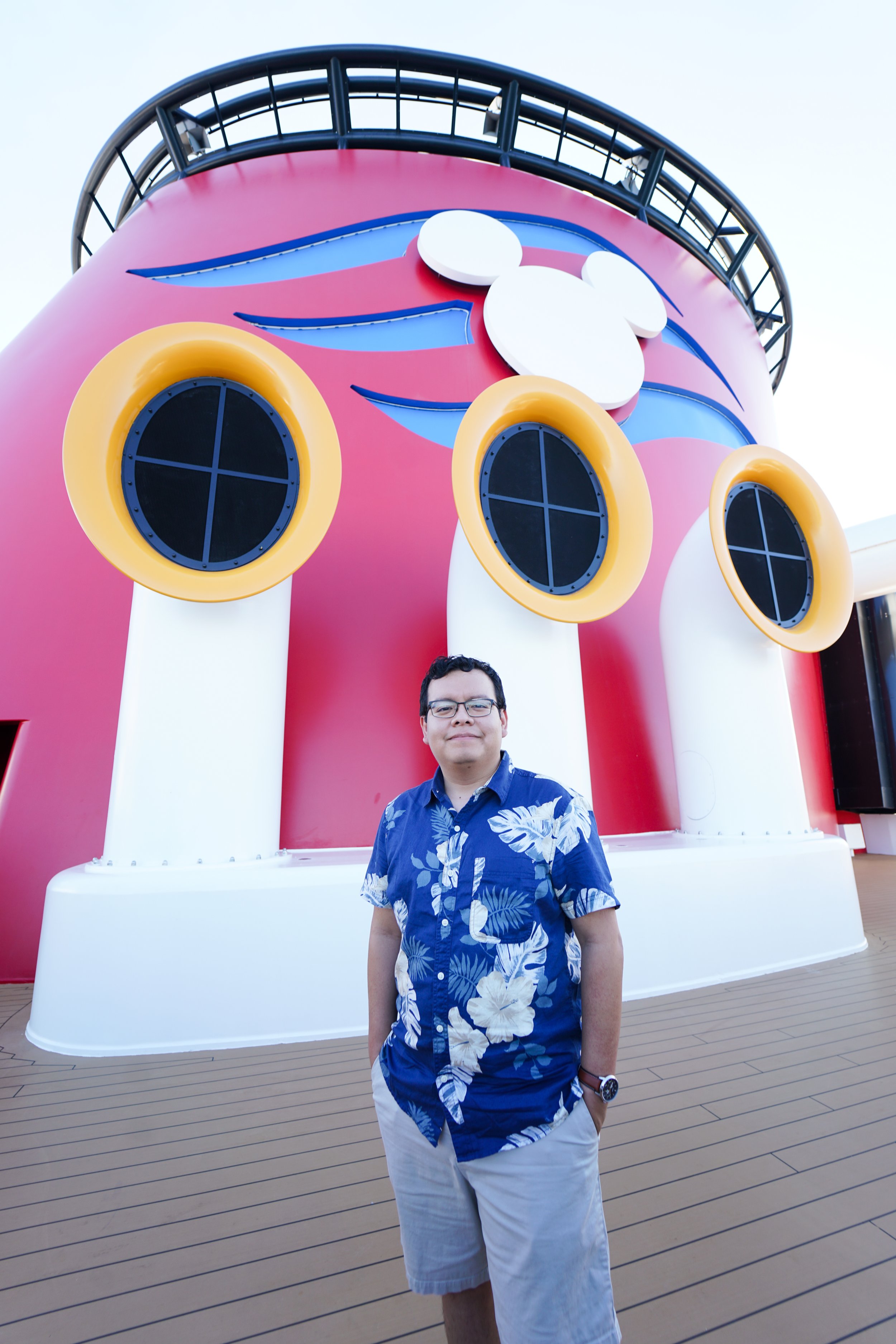 A man, the owner of AAR Photography, wearing glasses, a blue Hawaiian shirt, and beige shorts standing on a ship deck in front of a colorful, cartoonish pink and blue structure with Mickey Mouse shapes.