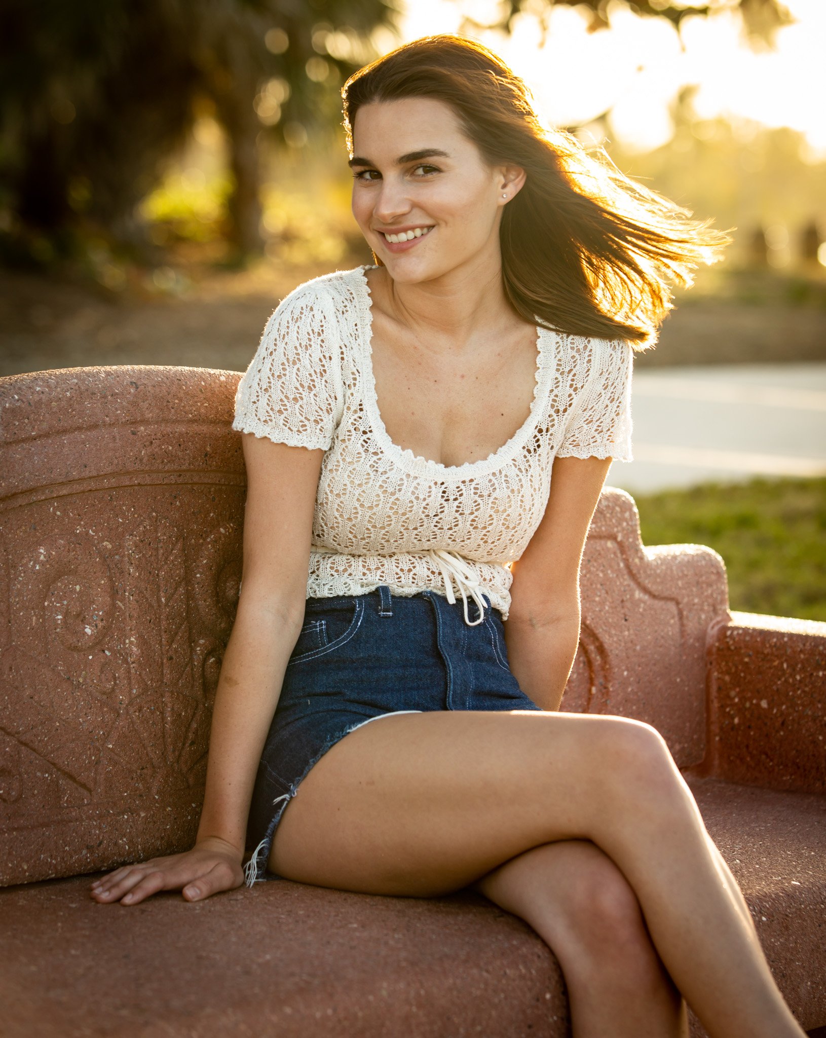 A young woman with brown hair sitting on a pinkish-brown stone bench outdoors during sunset, wearing a white crochet top and denim shorts, smiling at the camera with her hair blowing in the wind.