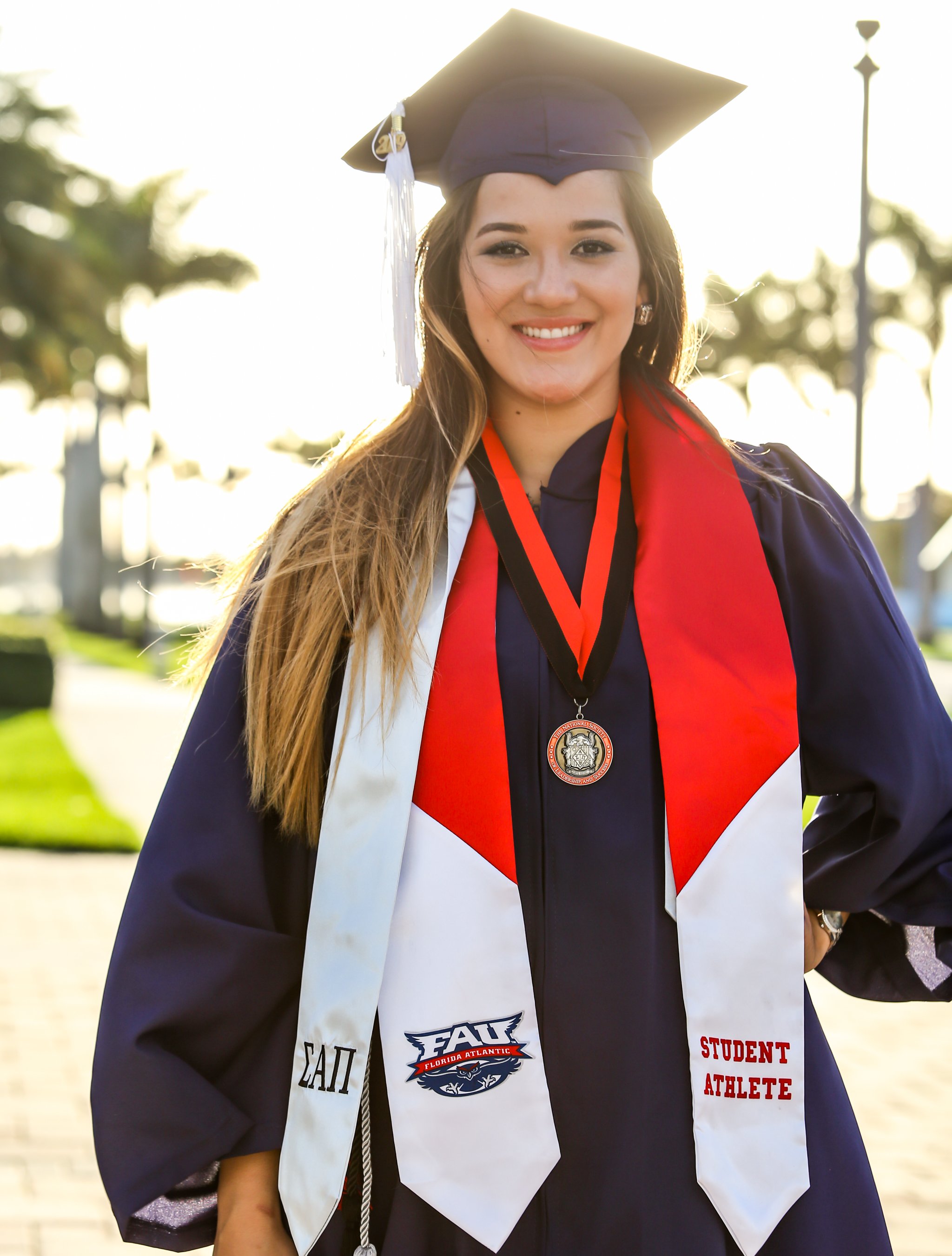 A young woman in a graduation cap and gown with a Florida Atlantic University Student Athlete stole smiles for the camera outdoors during sunset.