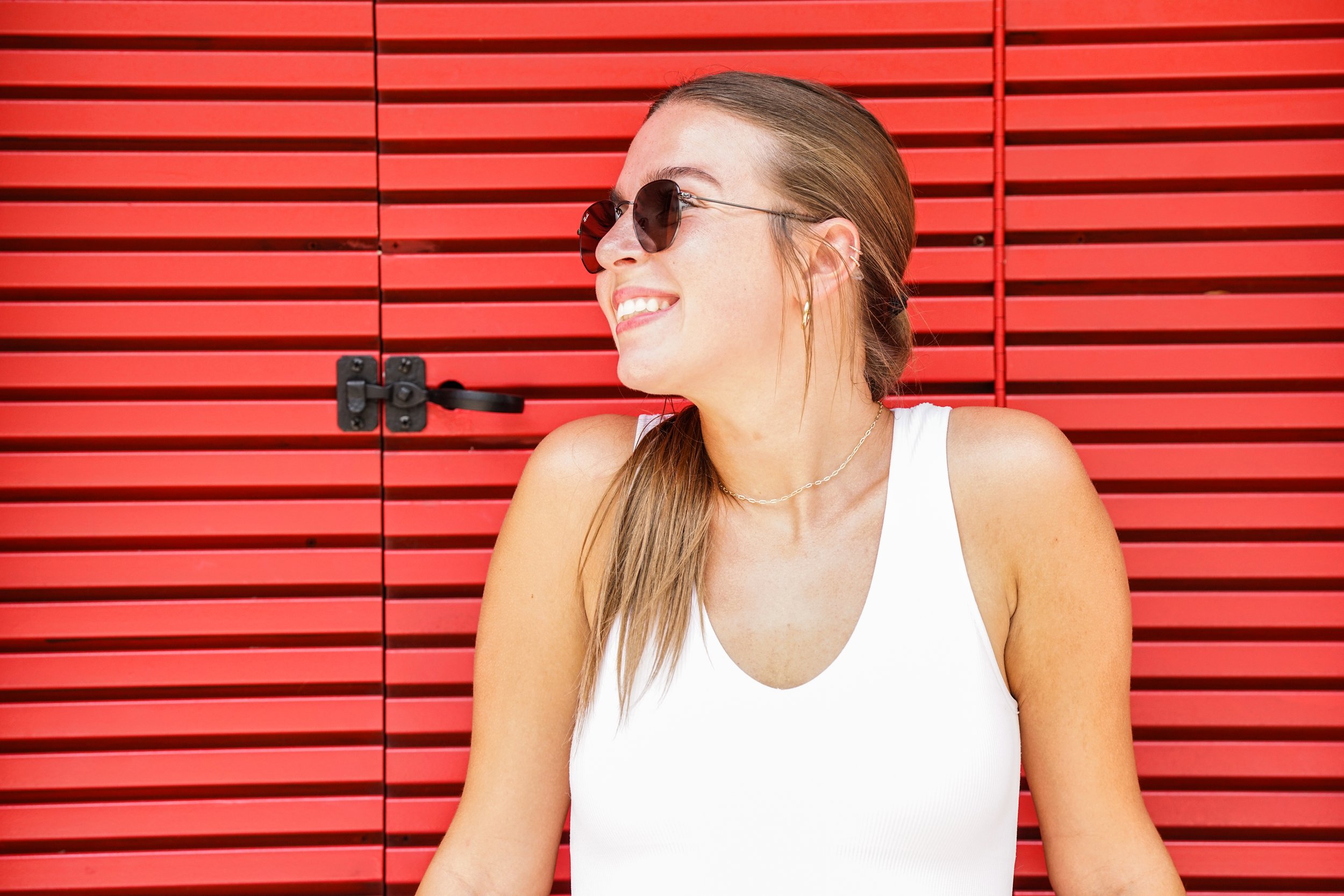 Side profile of a woman with long brown hair smiling, wearing dark sunglasses, gold hoop earrings, a gold necklace, and a white sleeveless top, standing in front of a red wooden wall.