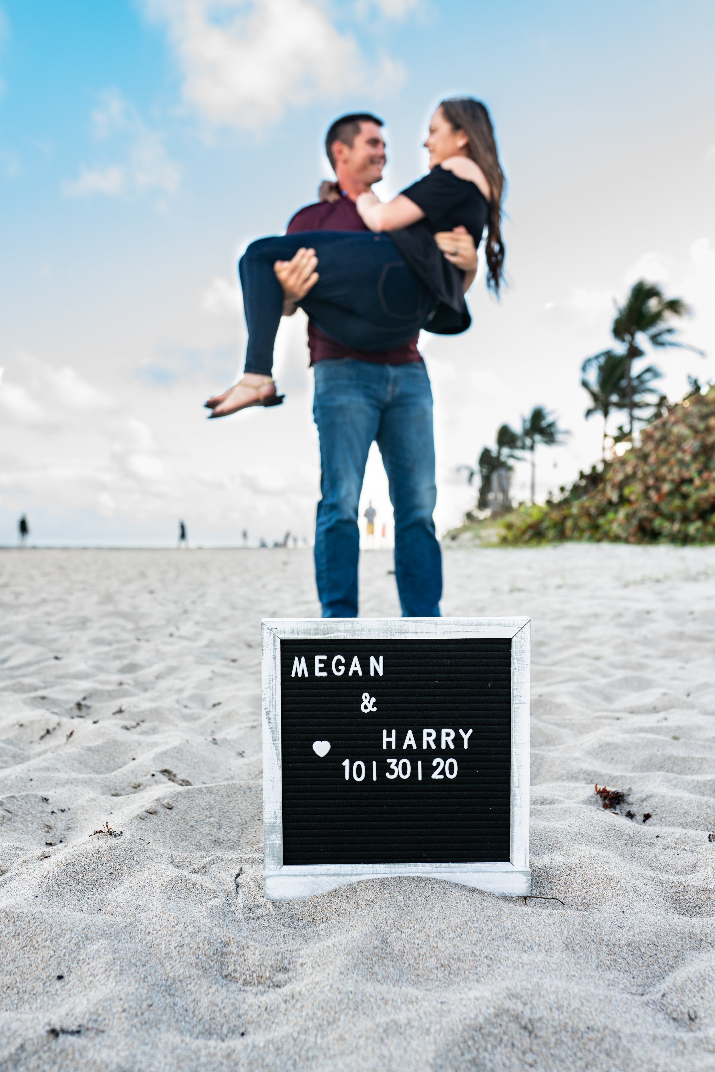 A man is holding a woman in his arms on a sandy beach with palm trees and a cloudy sky in the background, celebrating a wedding or engagement. A black letter board with white text reads "MEGAN & HARRY 10|30|20" in the foreground.